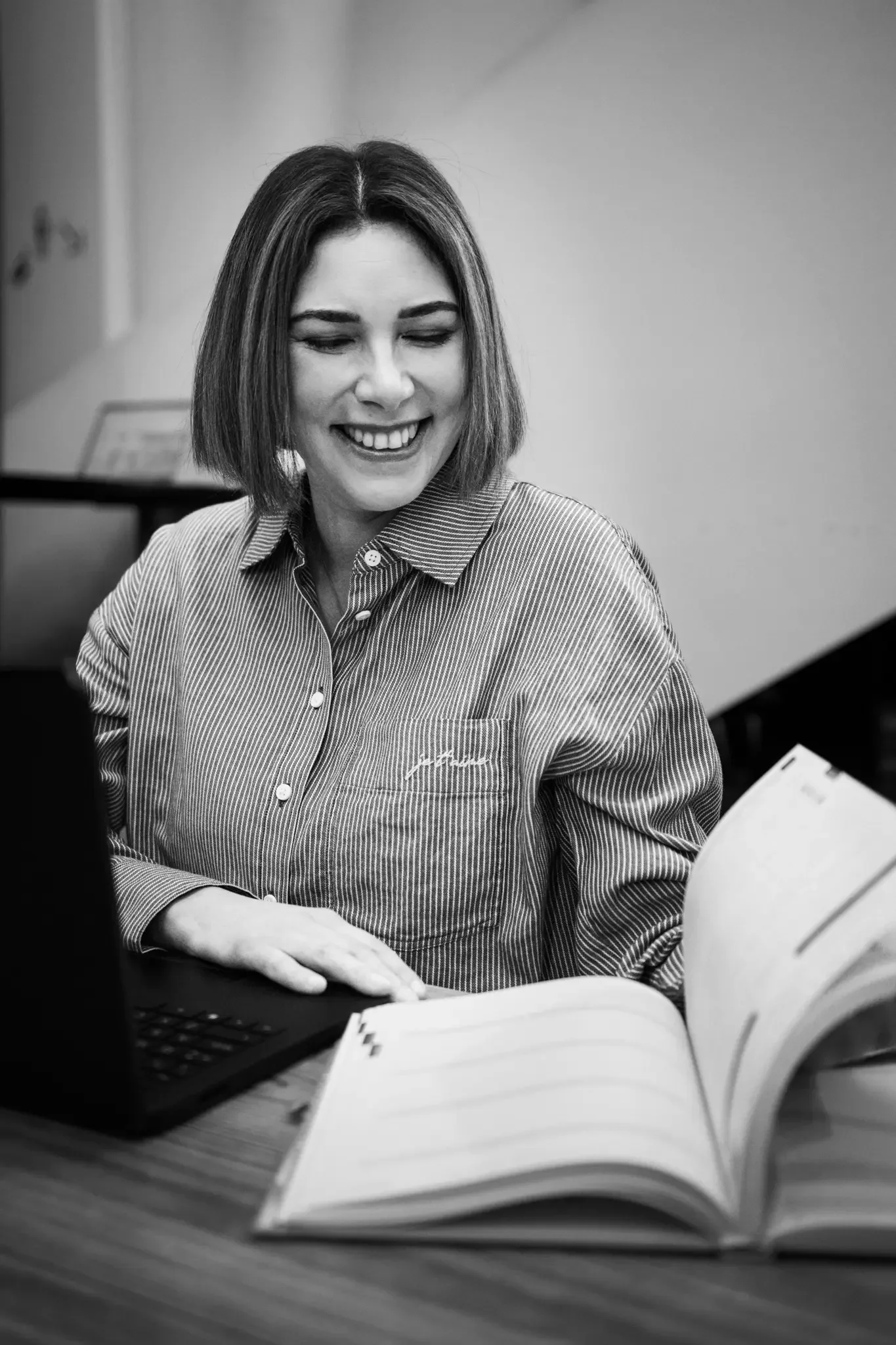 Black and white photo of a woman with shoulder-length hair, smiling while looking at an open book on a desk. She is wearing a striped button-up shirt and sitting in an office or classroom setting.