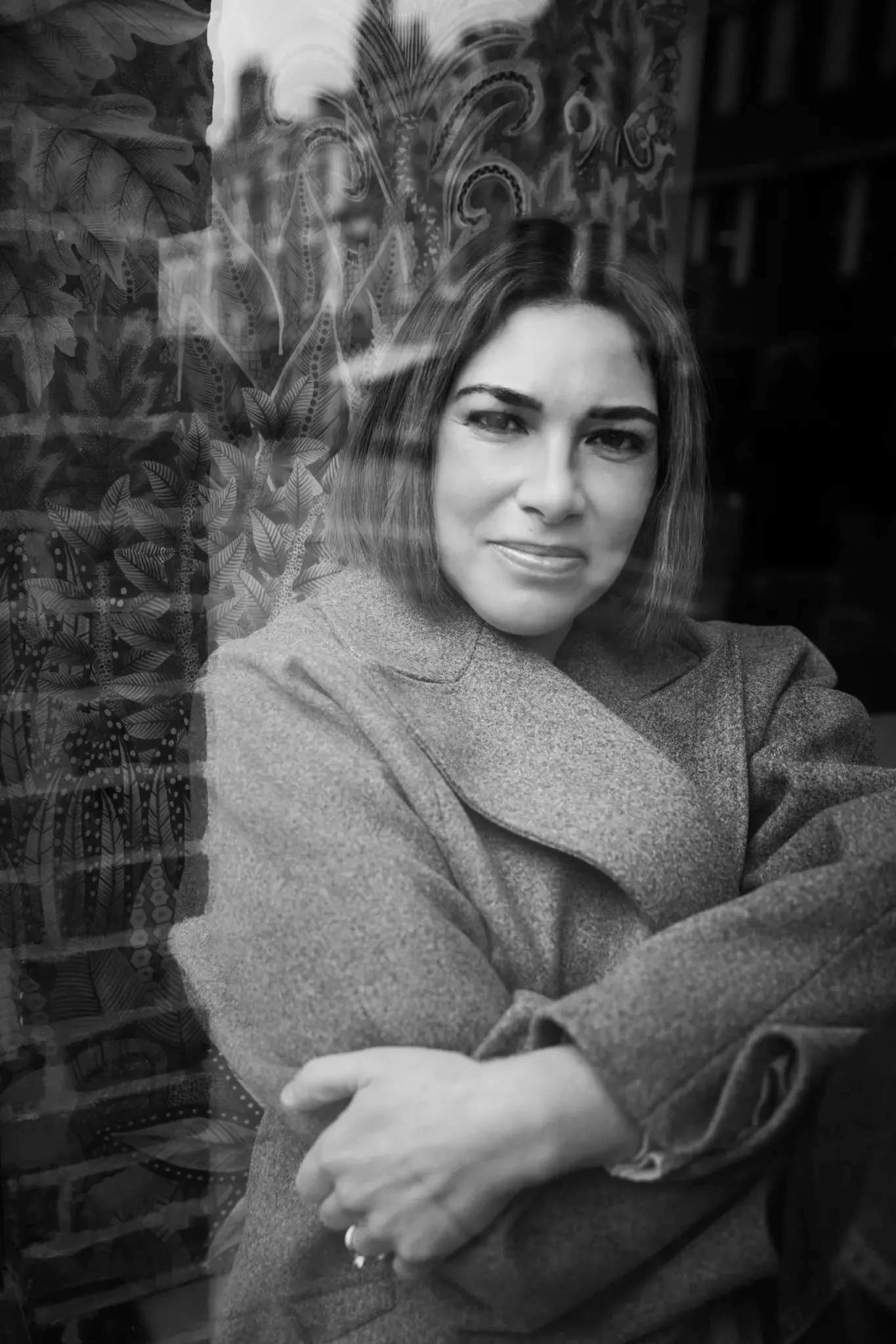 A woman with shoulder-length hair making a slight smile, sitting with arms crossed, seen through a window with decorative designs and foliage reflections.