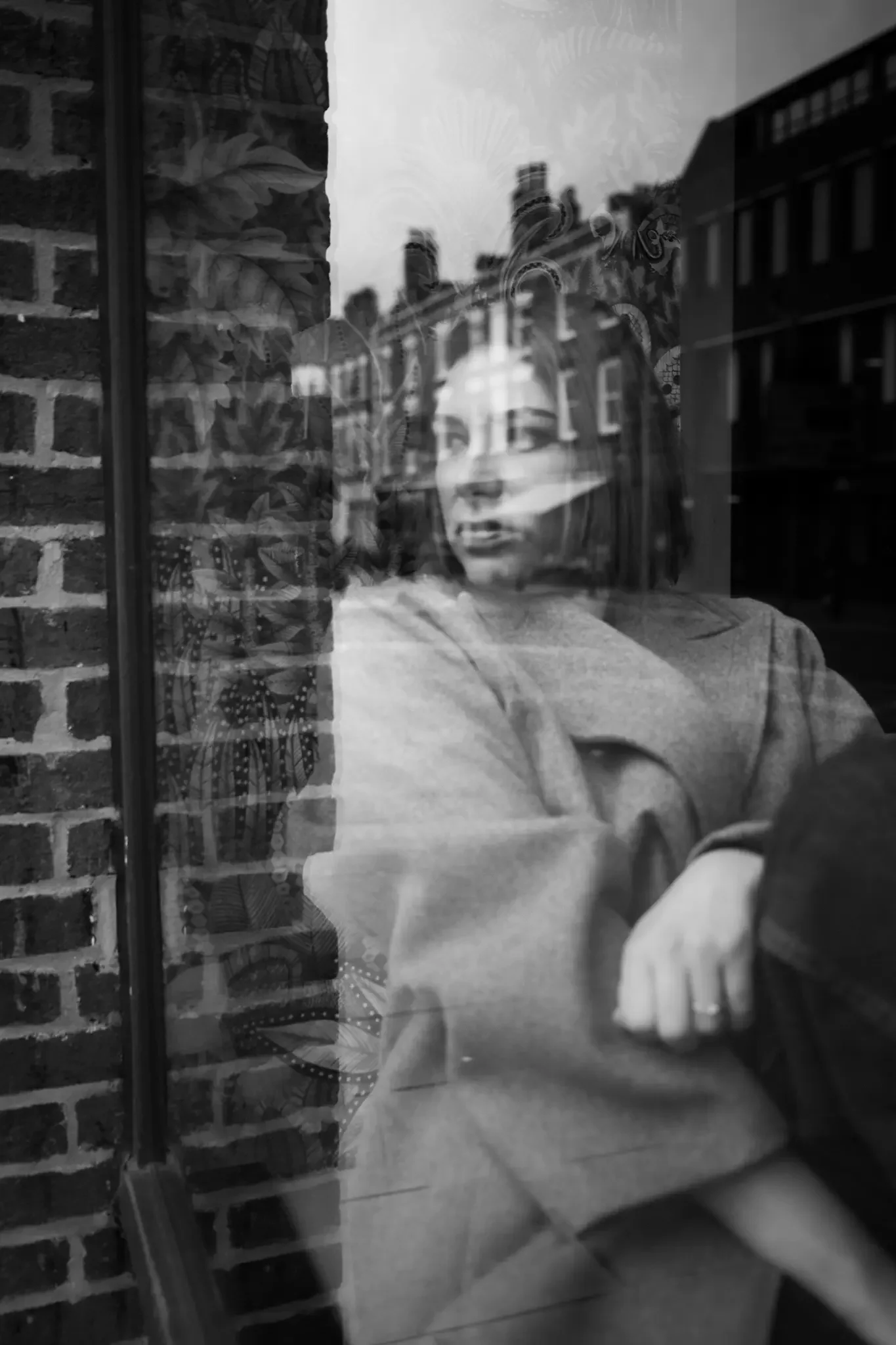 A woman looking out a window, with reflections of buildings and the street visible on the glass, creating a layered effect in the black and white photo.