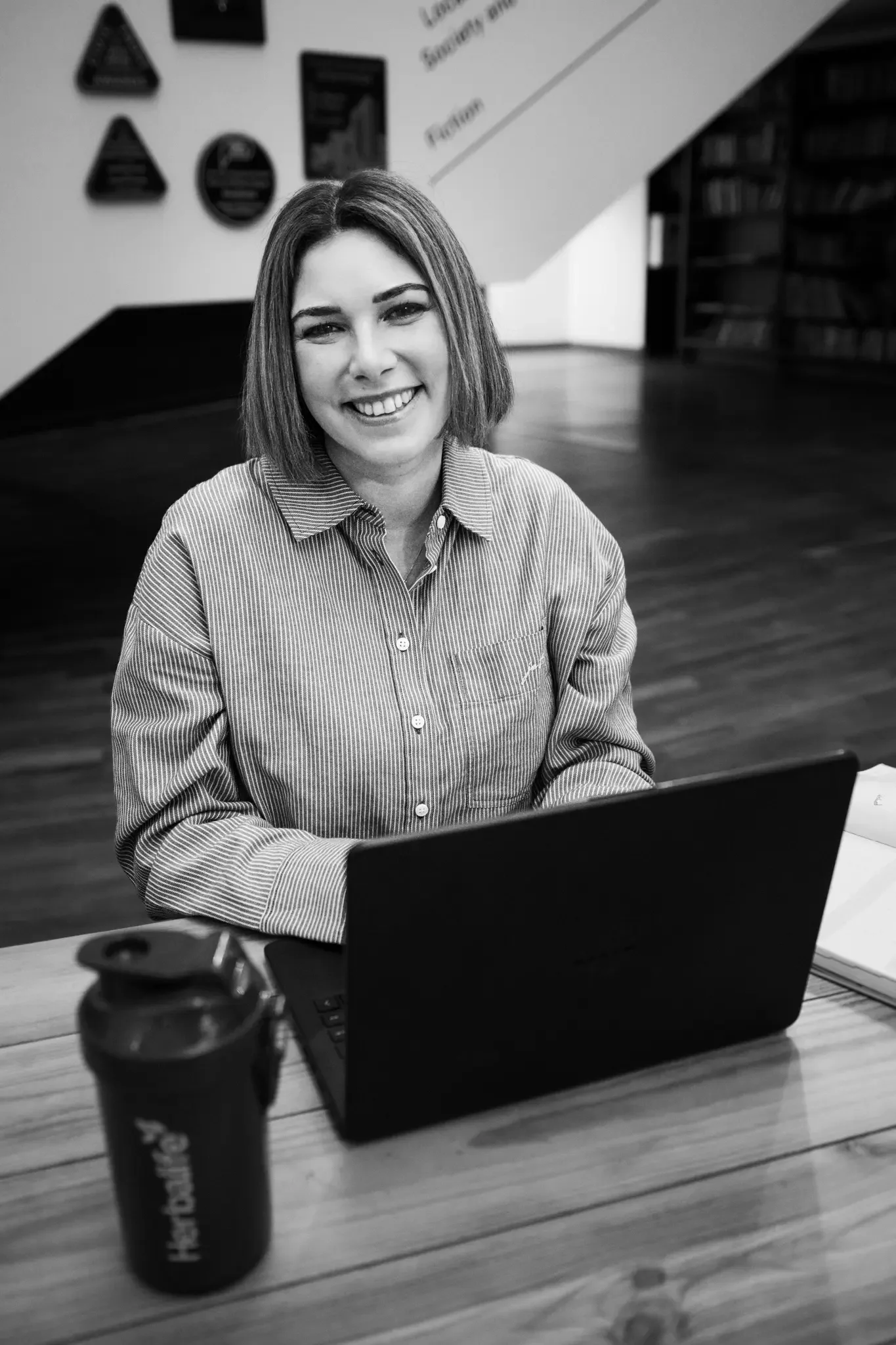 A smiling woman with shoulder-length hair, wearing a striped button-up shirt, sitting in front of a laptop on a wooden table, with a notebook and a reusable cup nearby, in a room with bookshelf and wall decor.