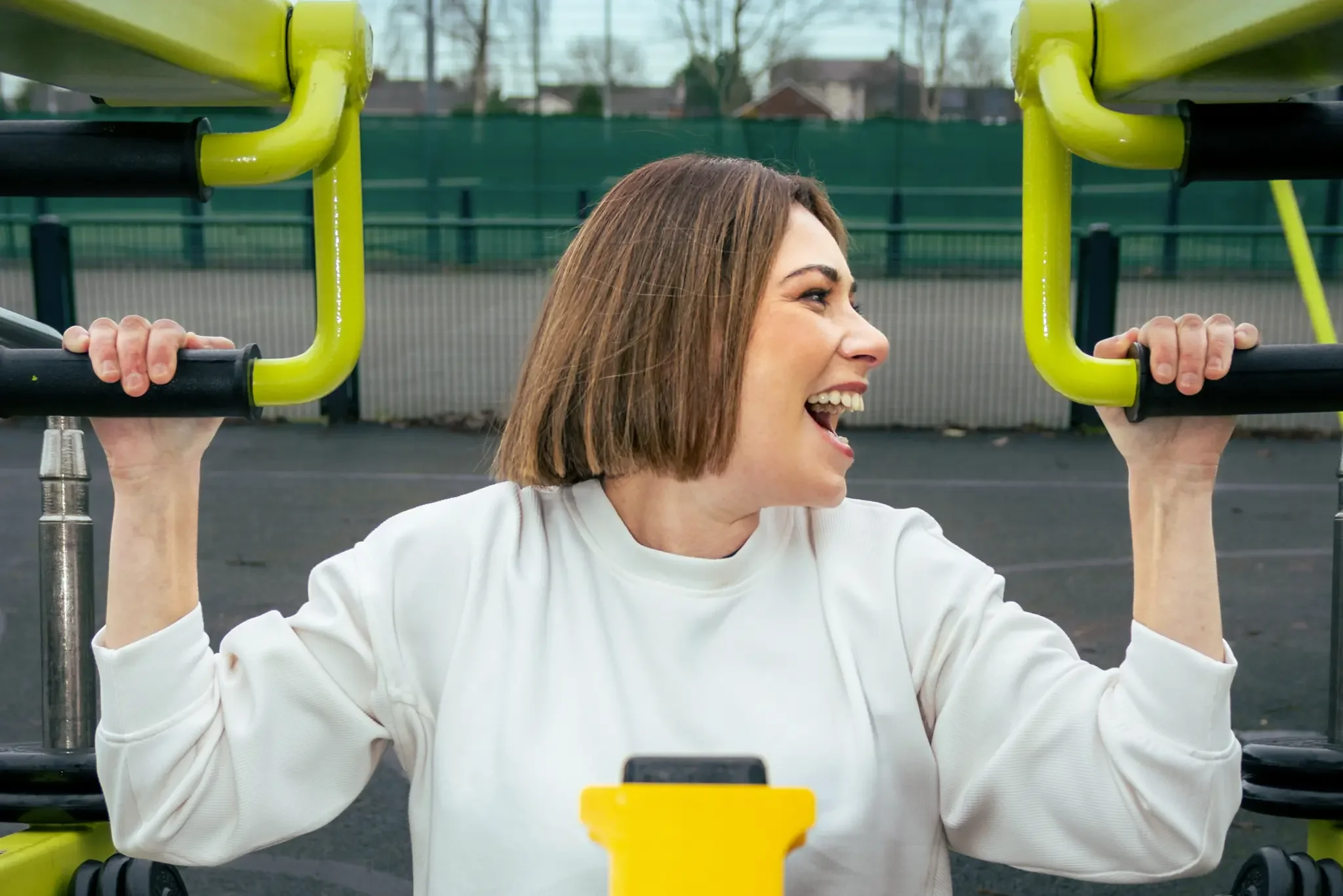 Woman with a bob haircut lifting a yellow and black gym machine at an outdoor fitness area, smiling energetically.