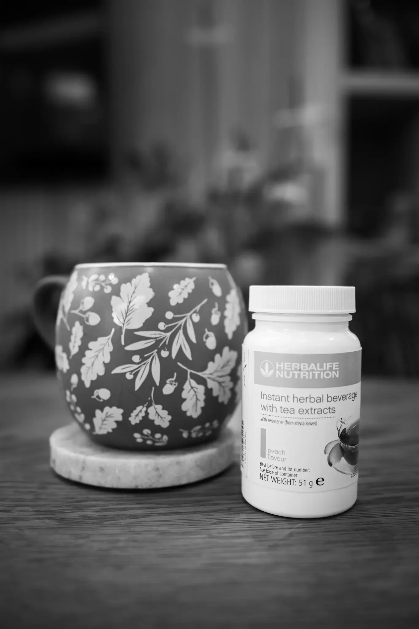 A decorated ceramic mug with a floral pattern placed on a coaster, next to a bottle of Herbalife nutritional supplement with tea extracts on a wooden surface.