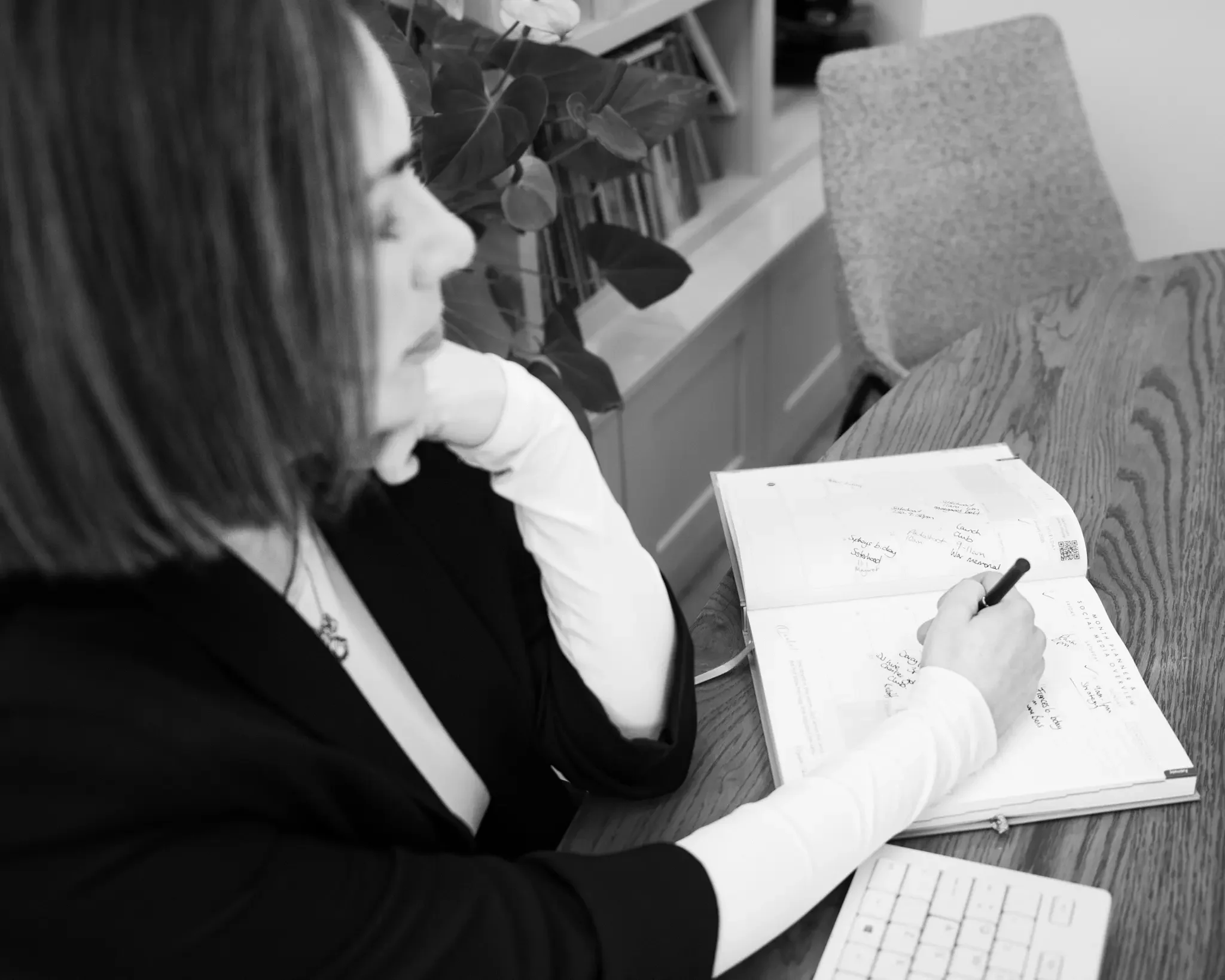 A woman with shoulder-length hair sitting at a wooden table, writing notes in a notebook, with a potted plant and shelf with books in the background.