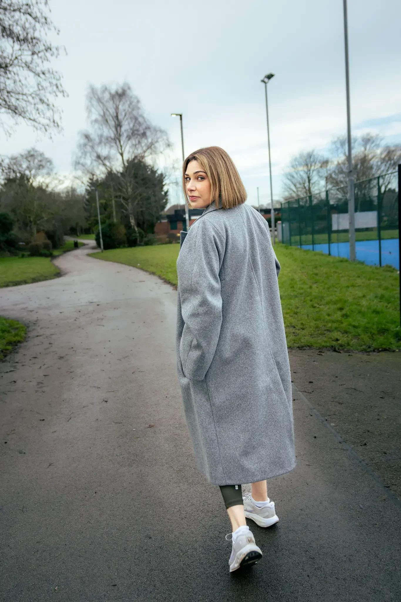 A woman in gray coat and sneakers walking on a park path, turning to look at the camera with trees and a tennis court in the background.
