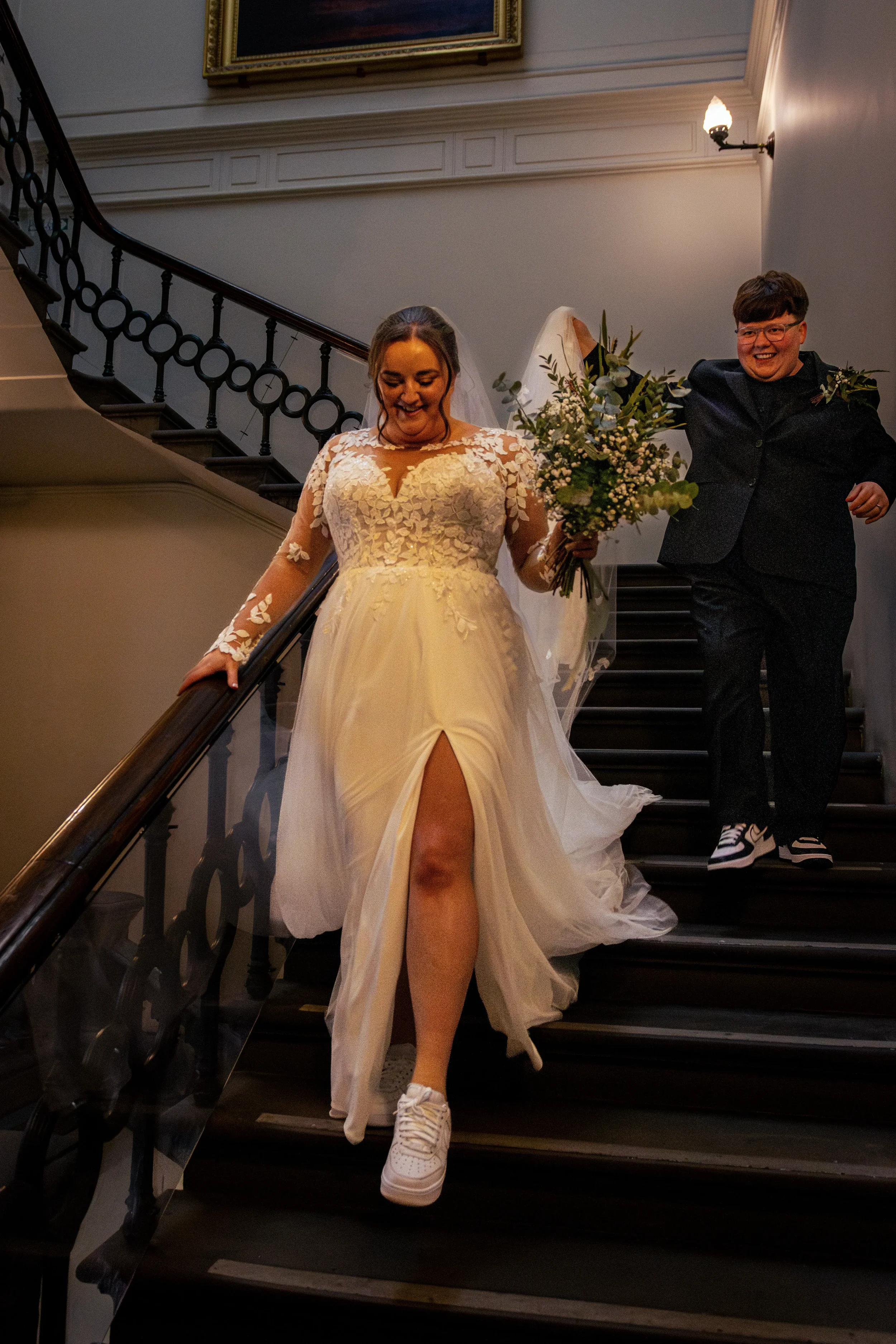 queer couple coming down stairs after ceremony. bride in suit carries bride in dress' veil.webp