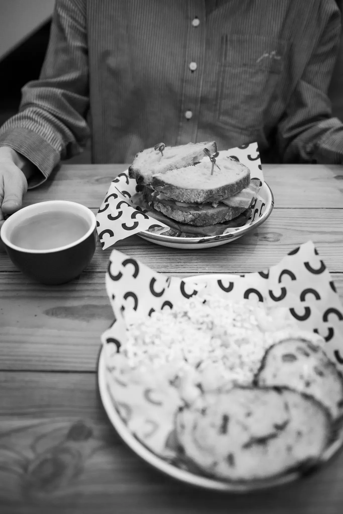A person sitting at a wooden table with a cup of coffee and two plates of food. The plate in the background has sandwiches held together with toothpicks. The plate in the foreground has cookies.