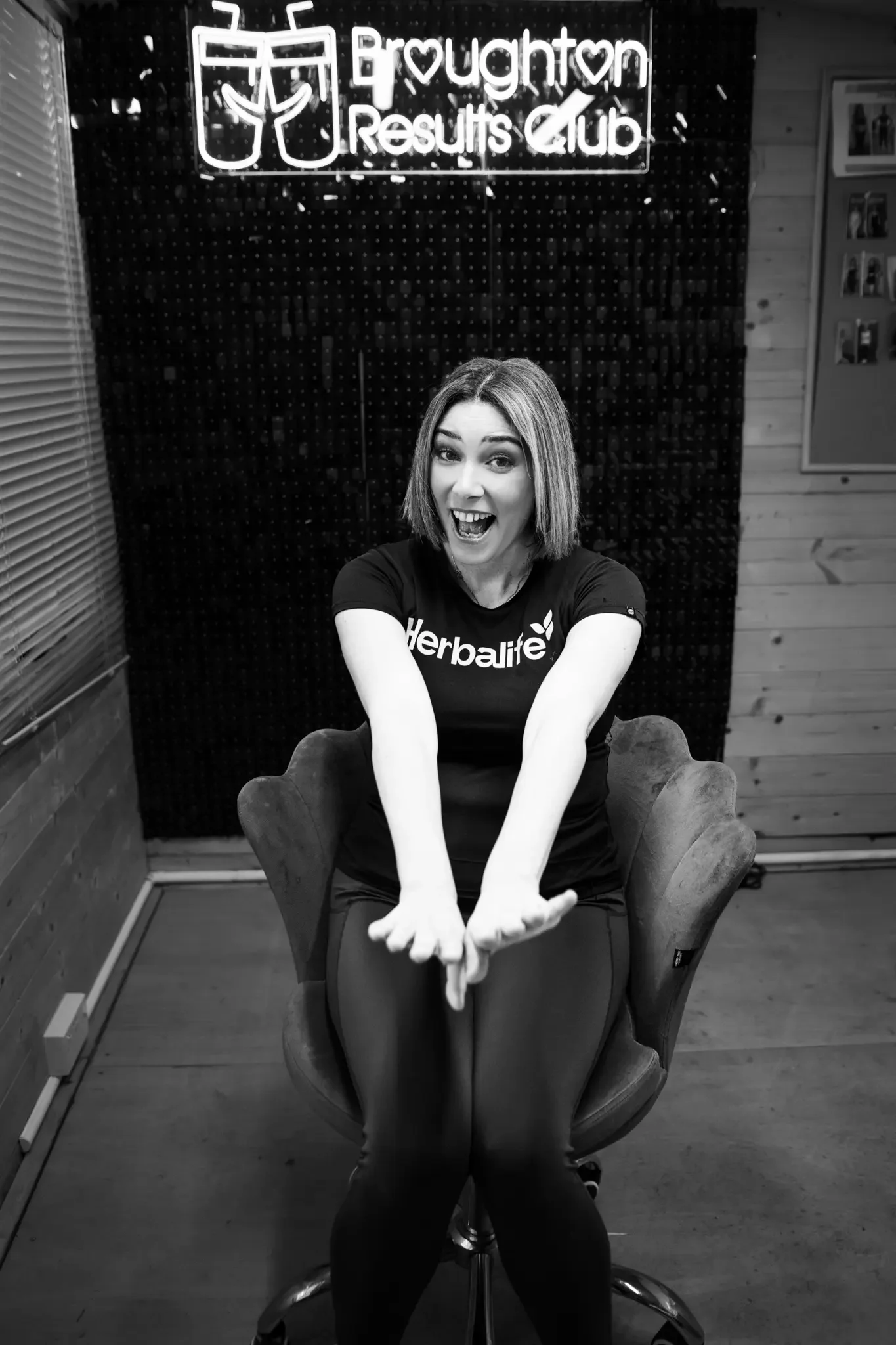 A woman sitting in a chair in front of a sign that says 'Broughton Results Club' with a graphic of two glasses with drinks. She is smiling and wearing a Herbalife t-shirt.