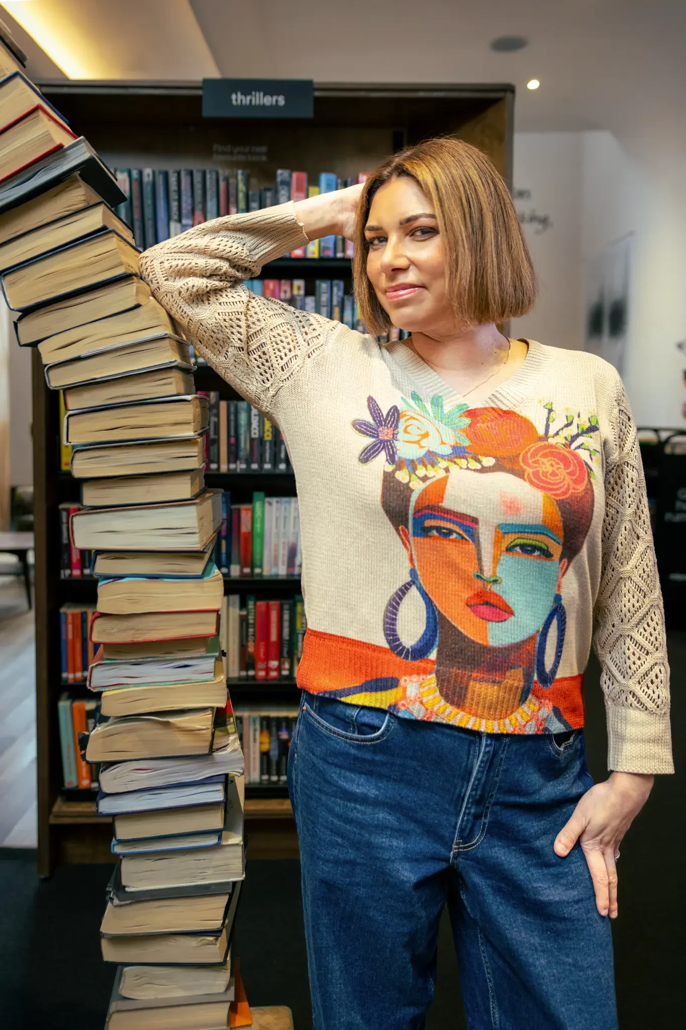 A woman with short brown hair and a colorful Frida Kahlo-themed sweater posing with a tall stack of books in a library.