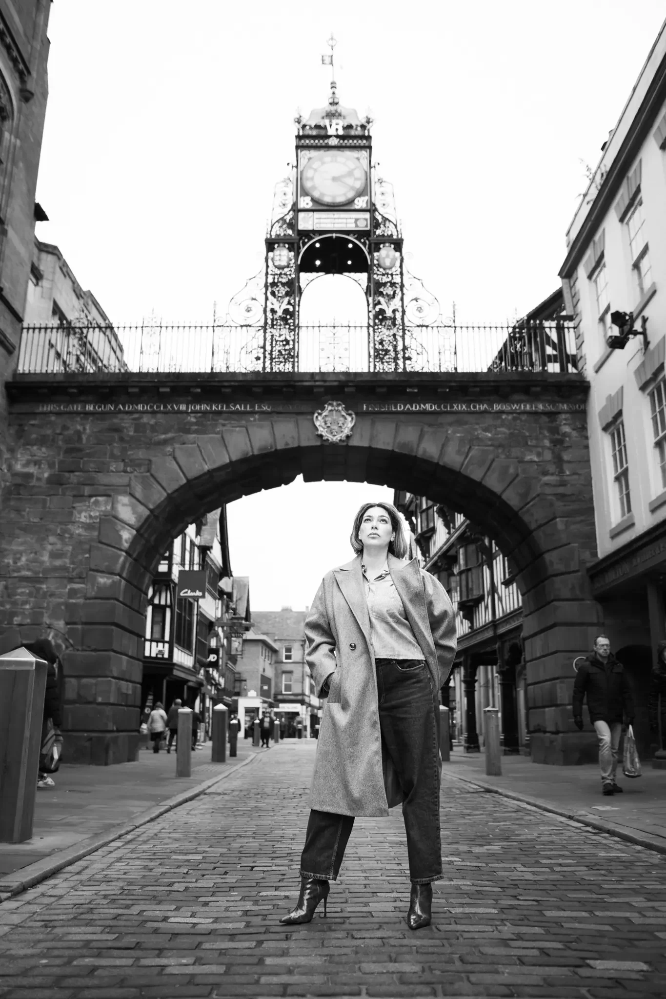 A woman stands confidently in the middle of a cobblestone street under a decorative clock tower, with buildings lining the street and other pedestrians in the background.