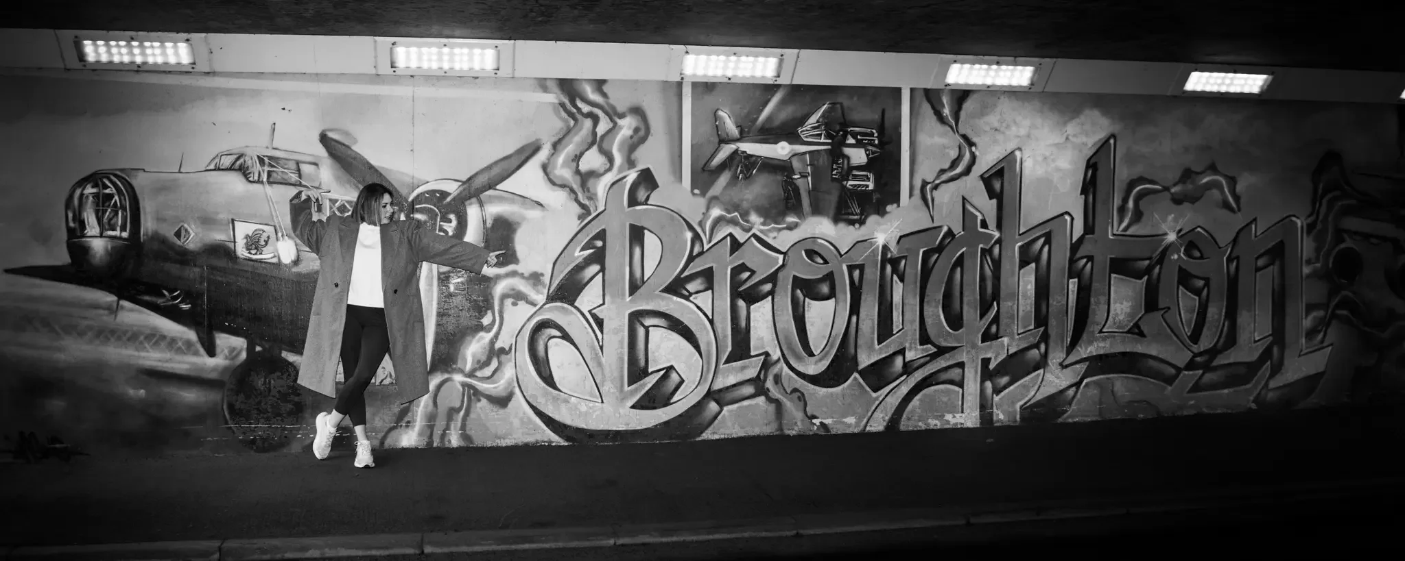 A woman in a long coat and sneakers pointing at a large graffiti mural of an airplane and the word "Brough" on a wall.