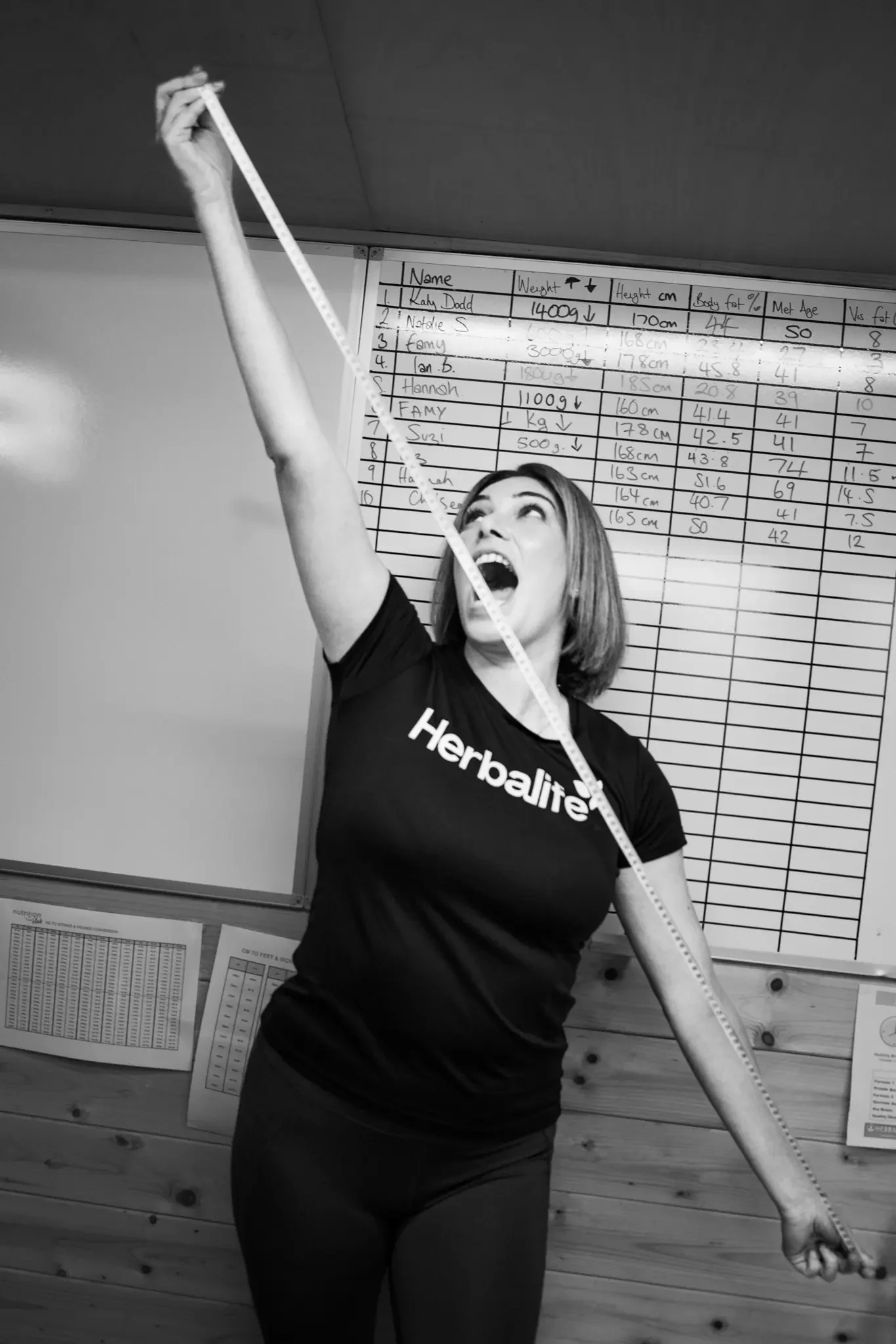 A woman wearing a Herbalife t-shirt is holding a measuring tape above her head, appearing excited and energetic, with a large chart of names and measurements behind her.