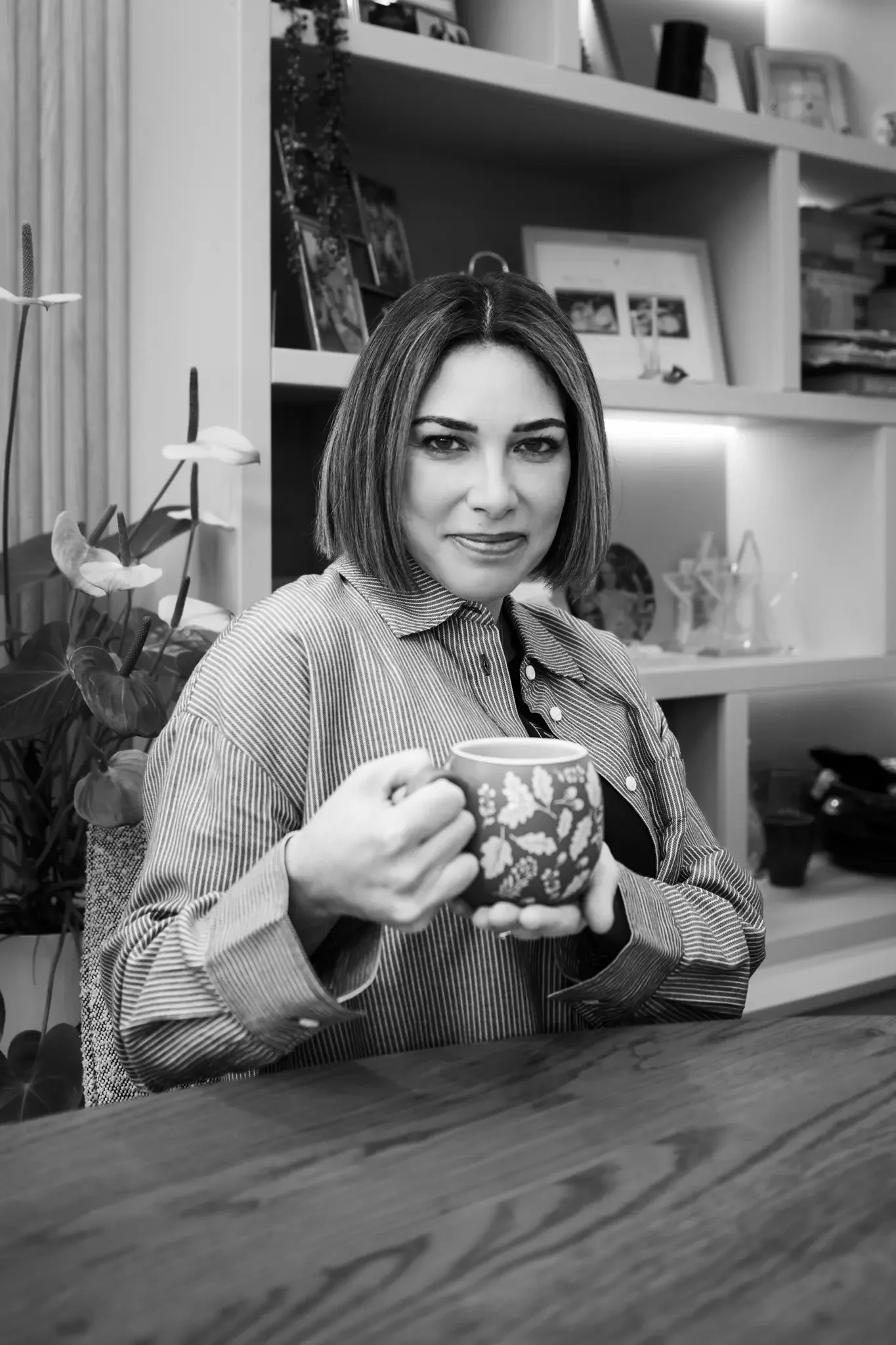 A woman with shoulder-length hair holding a decorative mug while sitting at a table, with shelves of framed photos and decorative items in the background.