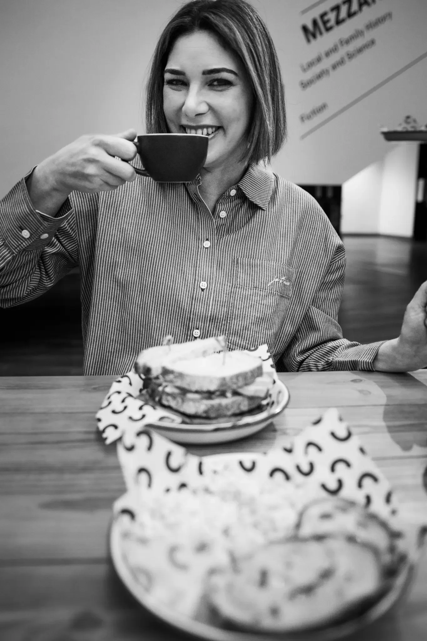 A woman with shoulder-length hair, wearing a striped button-up shirt, smiles and drinks from a cup while sitting at a table with a plate of toast and a small plate of cookies.