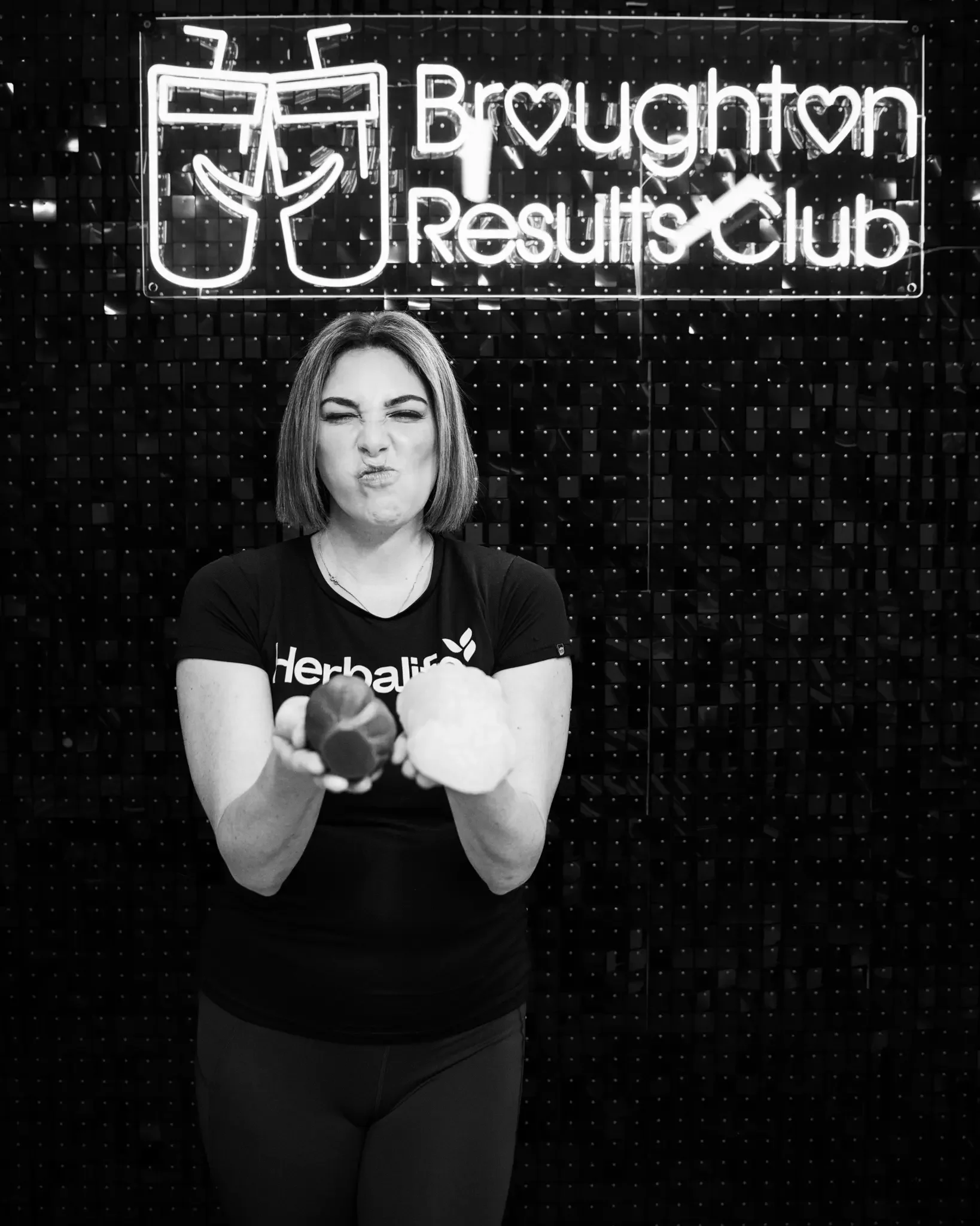 Woman holding a red and white cauliflower and a red bell pepper in front of a Broughton Results Club neon sign.
