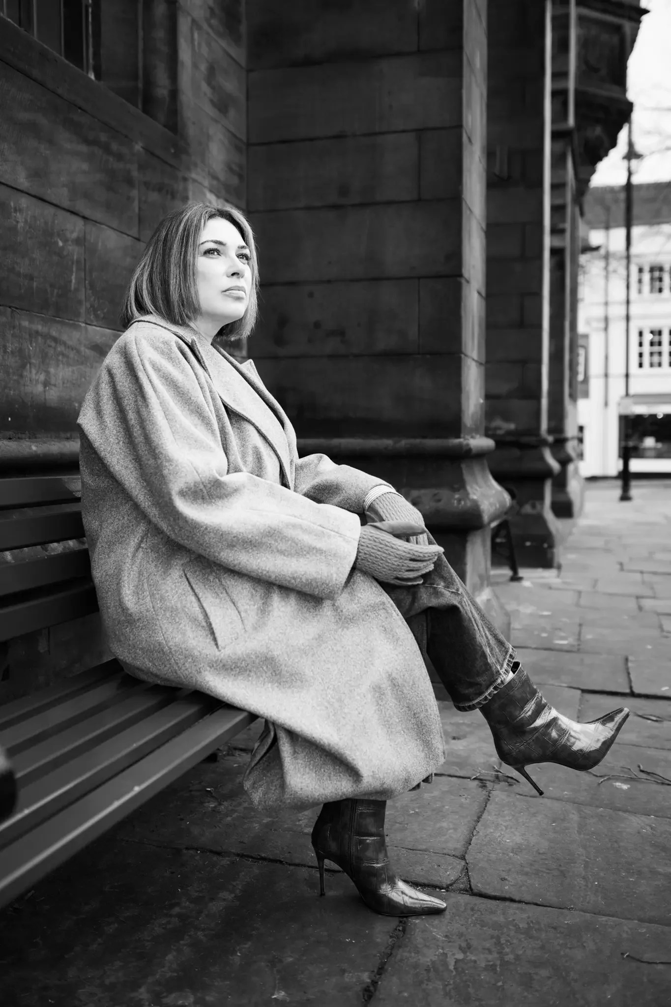 A woman with short hair sitting on a park bench wearing a long coat, gloves, and high-heeled boots in an urban area.