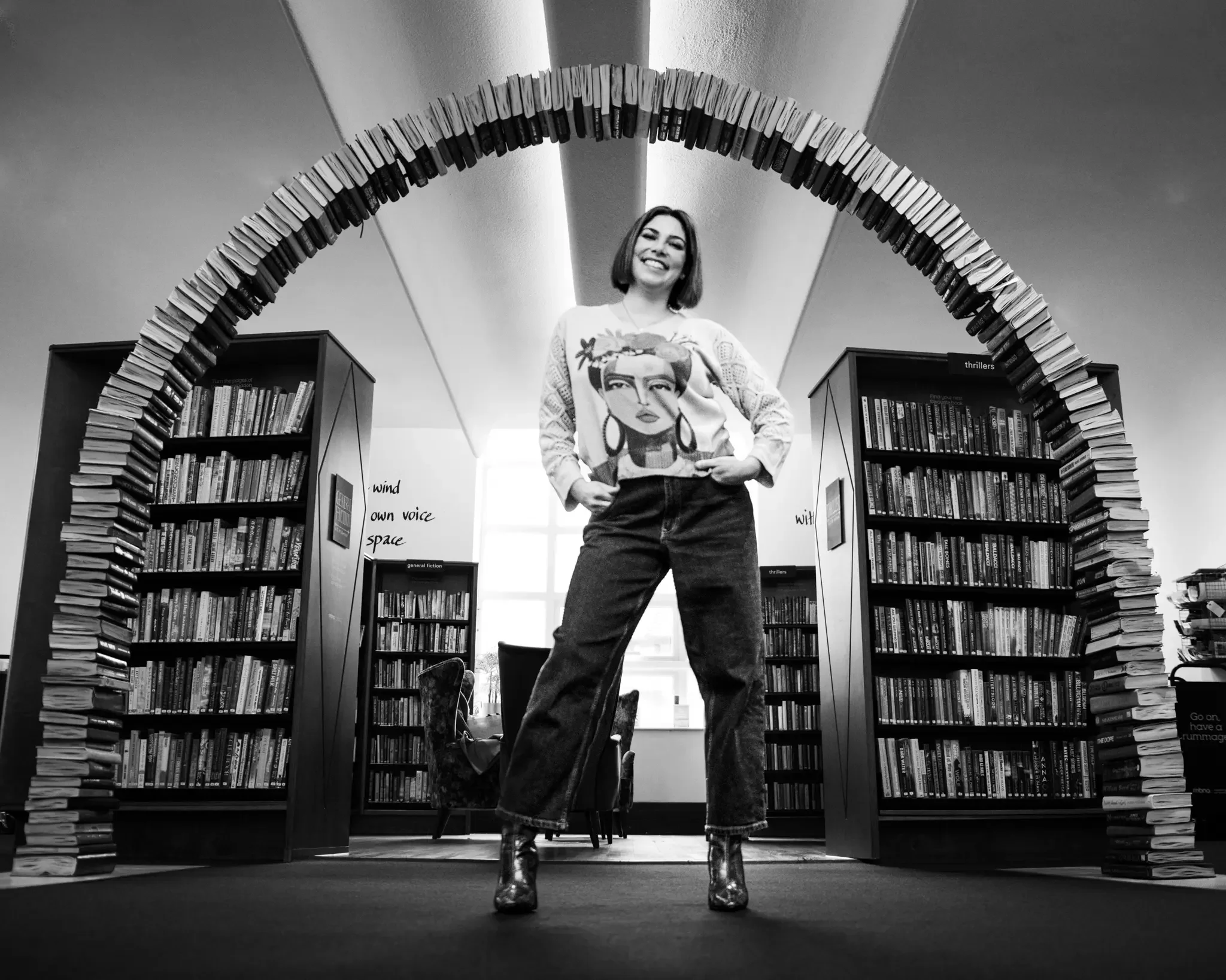 A woman standing in a library under an archway made of stacked books, smiling and looking at the camera, with bookshelves and words on the wall behind her.