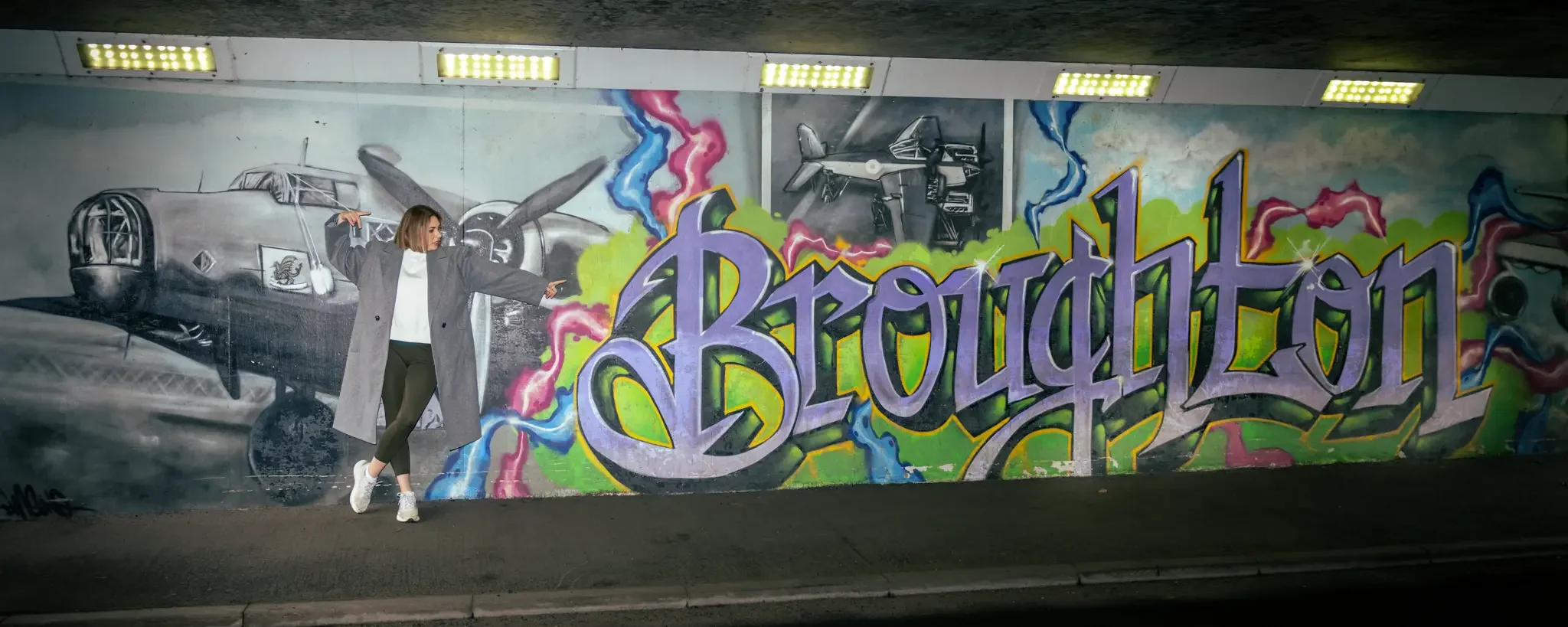 A woman standing in front of a colorful graffiti mural of a plane and the word 'Broughon' on a wall, with her arms extended as if pointing or posing.