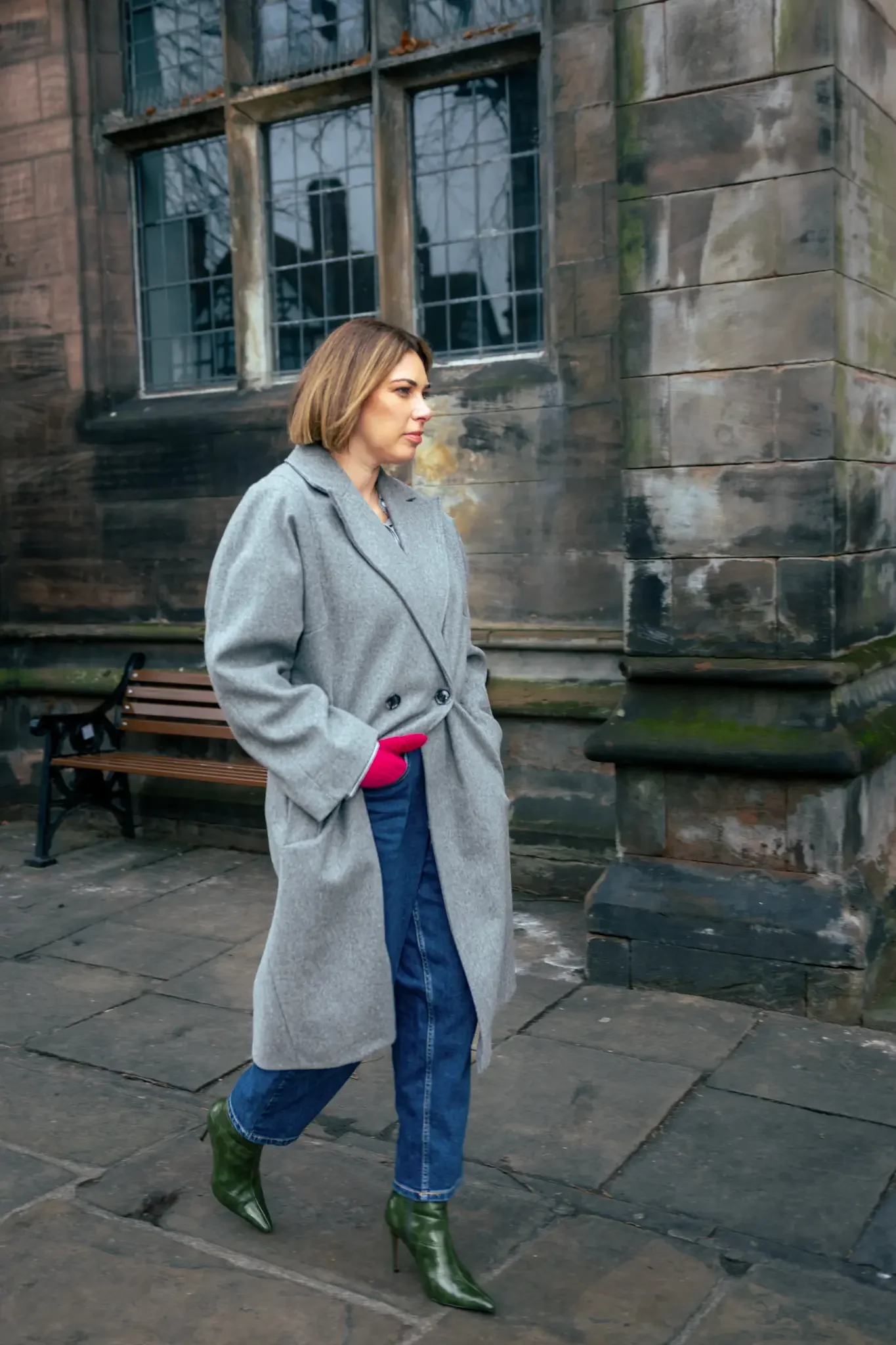 Woman walking on stone paved street outside, wearing a gray coat, blue jeans, green high-heeled boots, and pink gloves, with dark historic building walls and windows in the background.