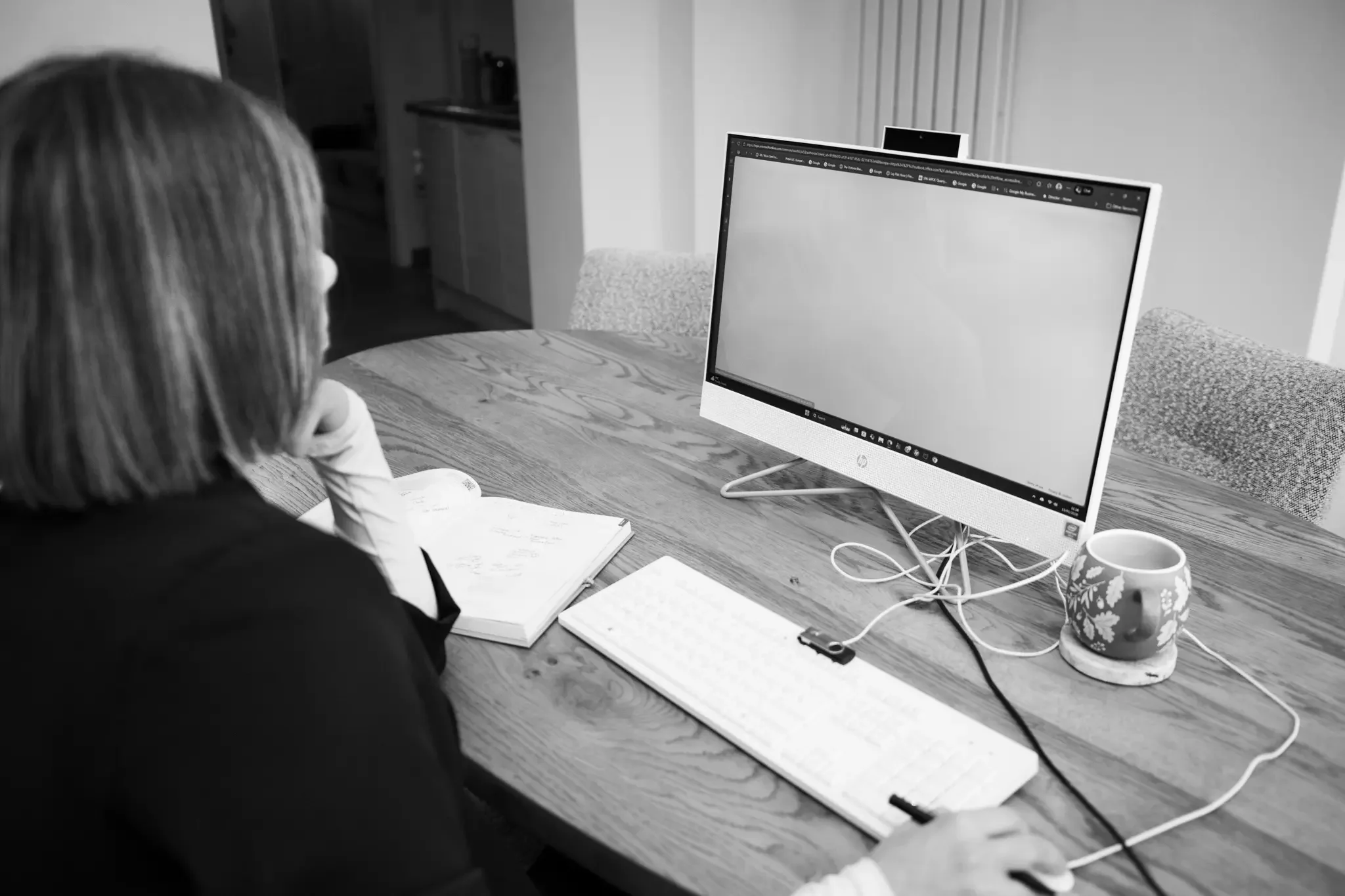 A woman sitting at a wooden table working on a computer with a notebook and mug nearby.