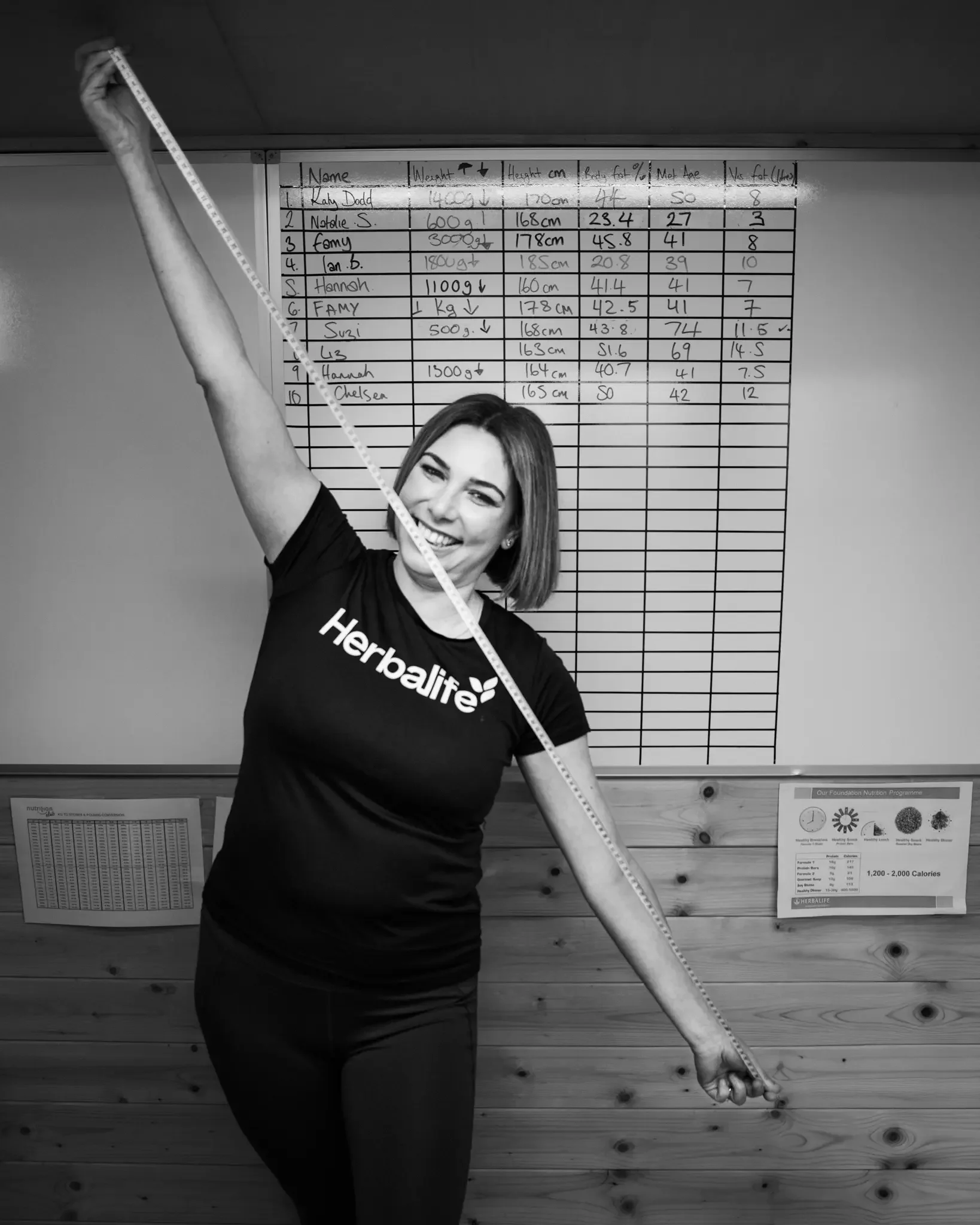 A smiling woman in a Herbalife T-shirt holding a measuring tape in a fitness or nutrition center, with a large chart on the wall behind her.