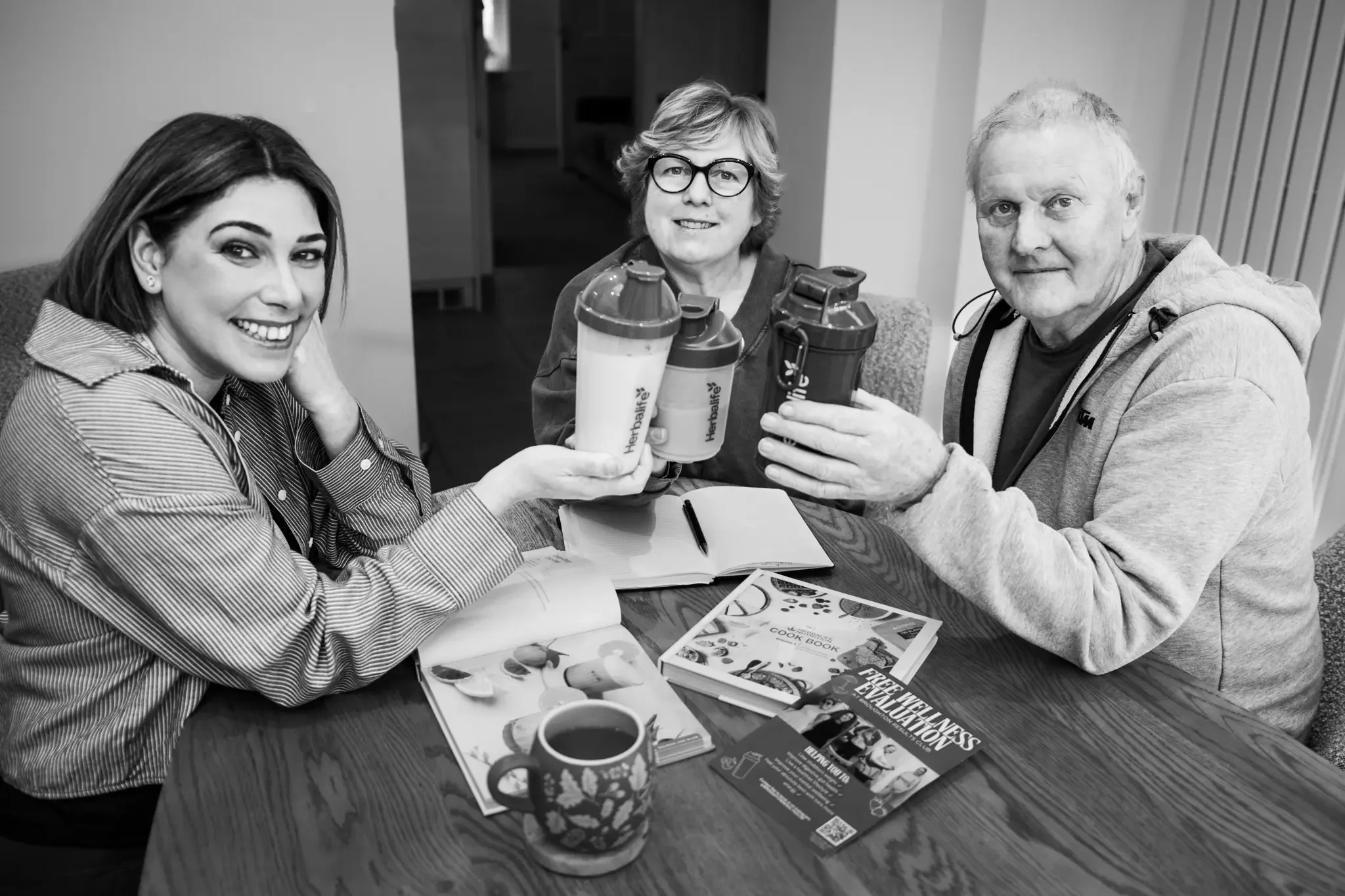Three people sitting at a table, holding up water bottles, smiling, with notebooks and flyers in front of them.