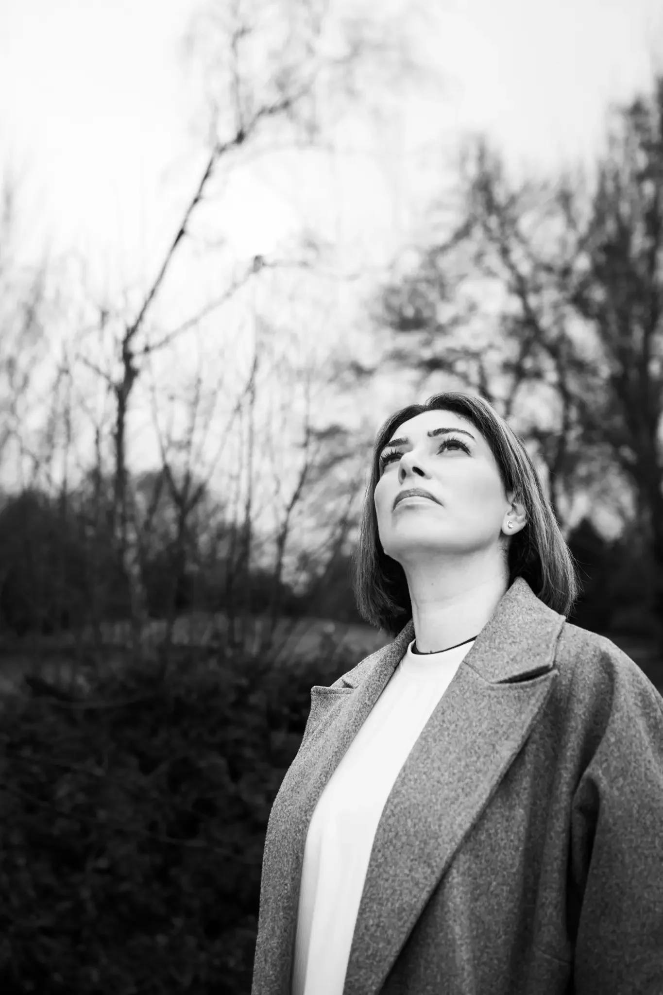 A black and white photo of a woman outdoors, looking upward with a contemplative expression, surrounded by trees with bare branches.