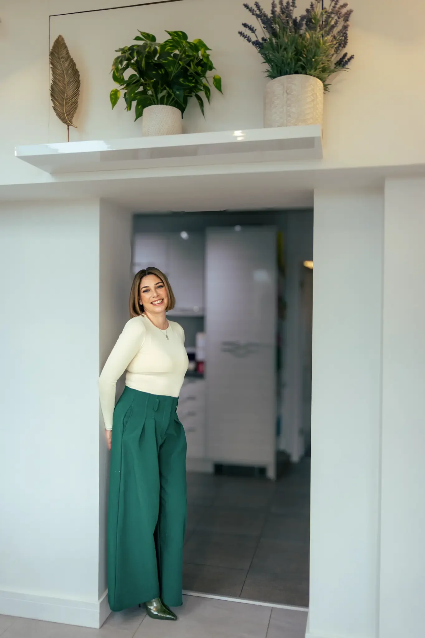A woman standing in a doorway smiling, wearing a white long sleeve top and green wide-leg pants, in a modern kitchen interior with white walls and decorative plants on a shelf above.