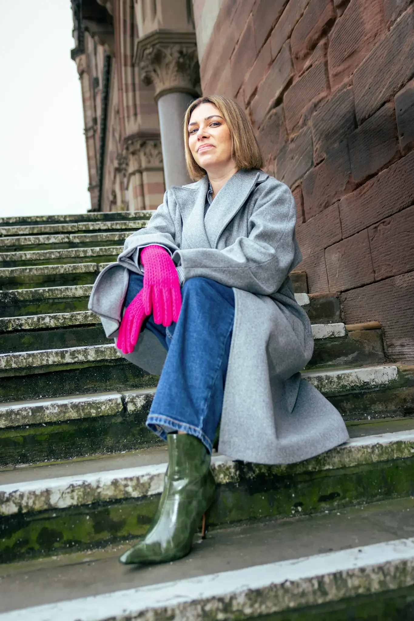 A woman sitting on moss-covered outdoor stairs, wearing a gray coat, blue jeans, green high-heeled boots, and bright pink gloves, with a brick building in the background.