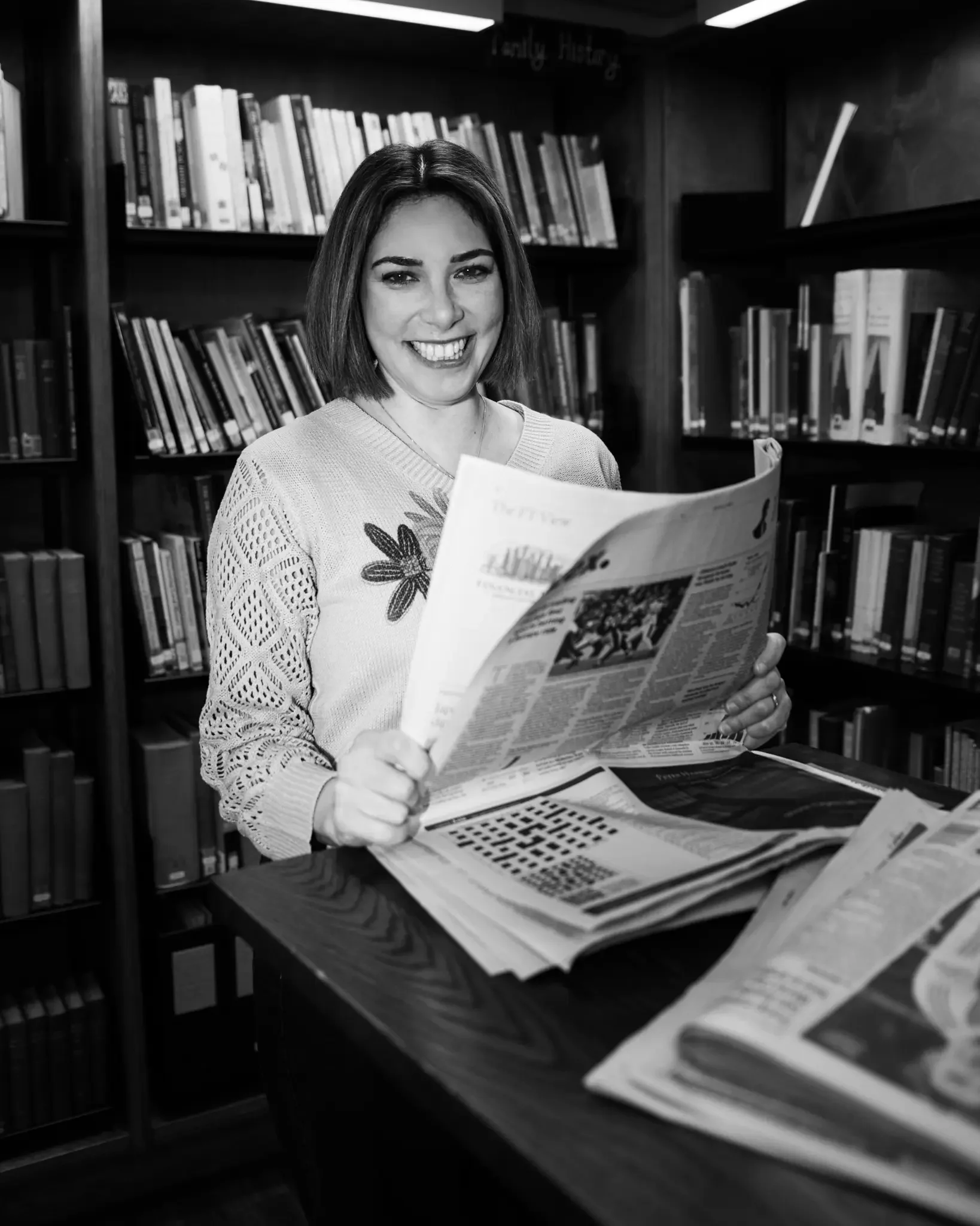 A smiling woman with shoulder-length hair reading a newspaper in a library, with shelves of books in the background.