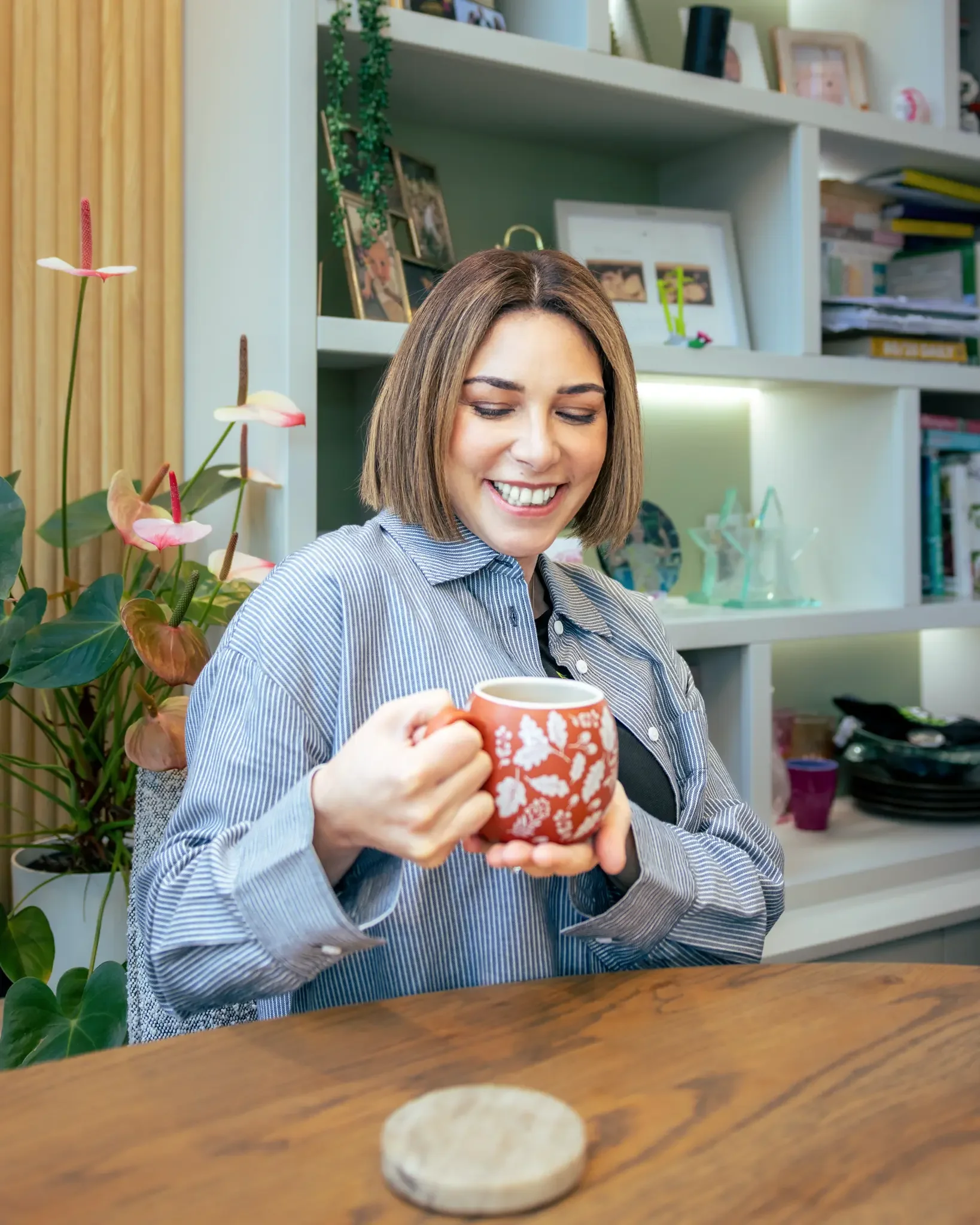 A woman with short brown hair smiling, holding a red mug with white floral pattern, sitting at a wooden table in a cozy home setting with plants and books in the background.