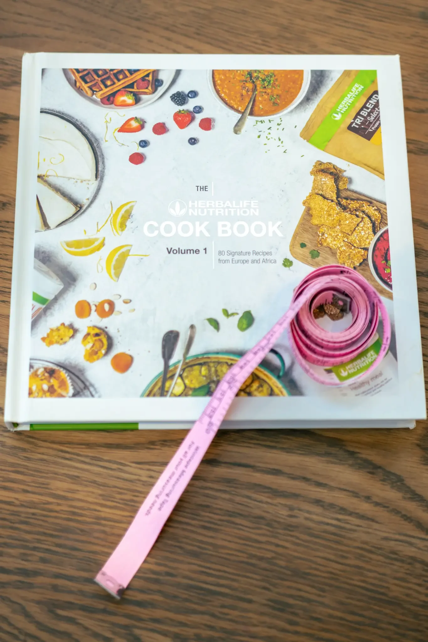 The Herbalife Nutrition Cook Book Volume 1 on a wooden surface, with a pink measuring tape placed on top of it.