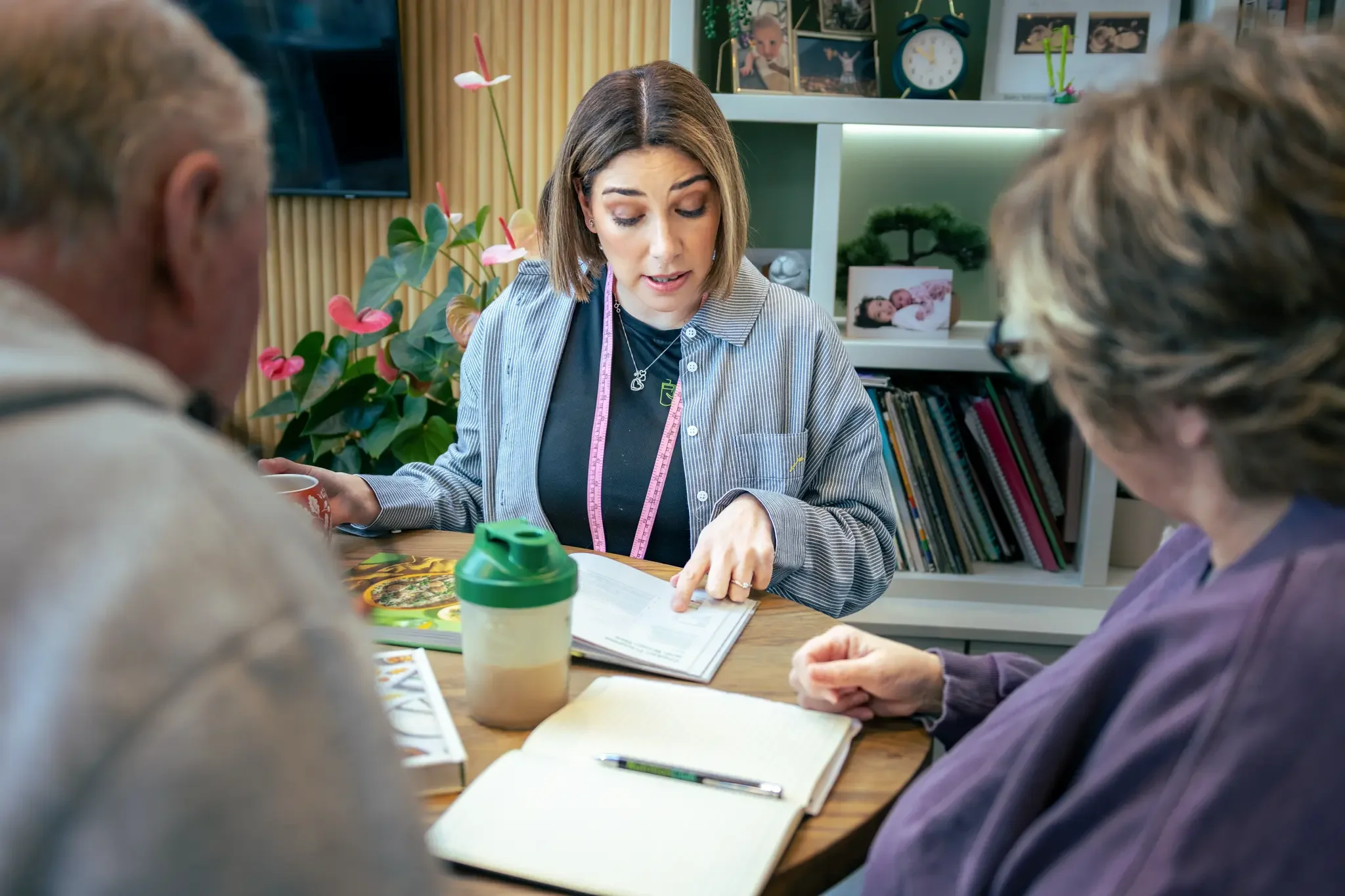A woman with shoulder-length brown hair showing documents to two elderly women at a table, with books, a coffee cup, and a notebook in front of her.