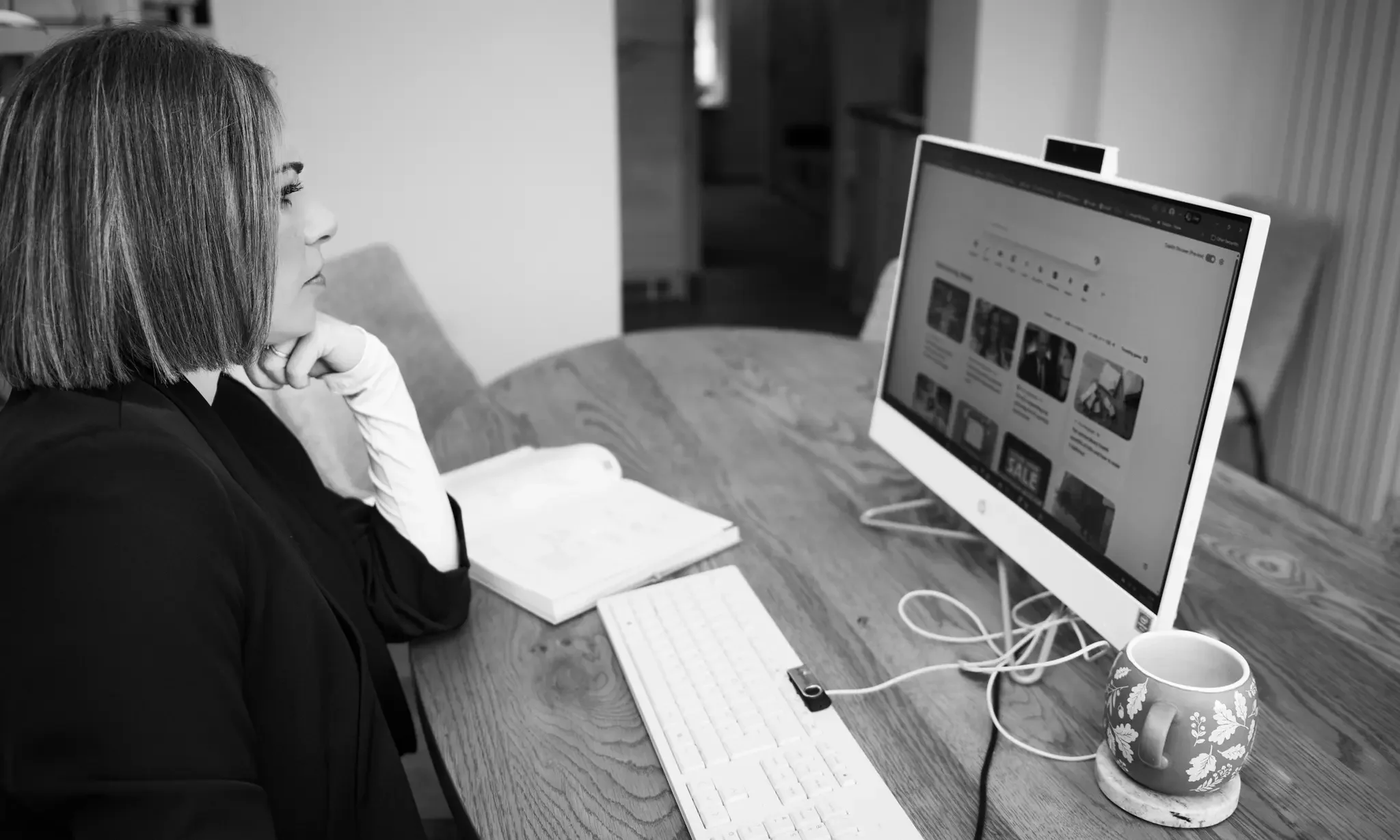 A woman with shoulder-length hair, wearing a dark blazer and white gloves, sitting at a wooden table in front of a desktop computer, looking at the screen. The desk has a notebook, a keyboard, a mug with floral design, and cables connected to the com