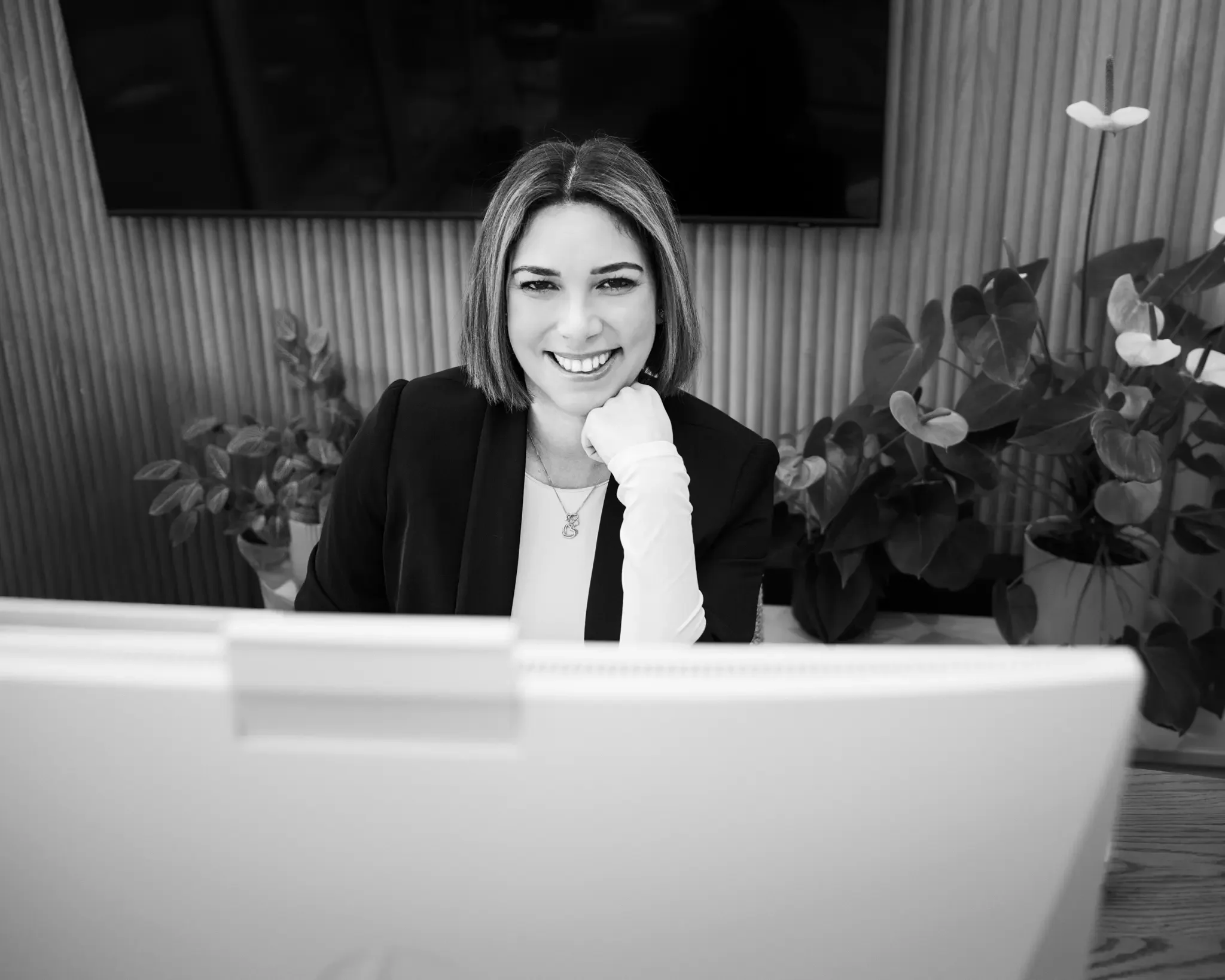 A woman smiling at her laptop in an office setting, with plants and a flat screen TV in the background.