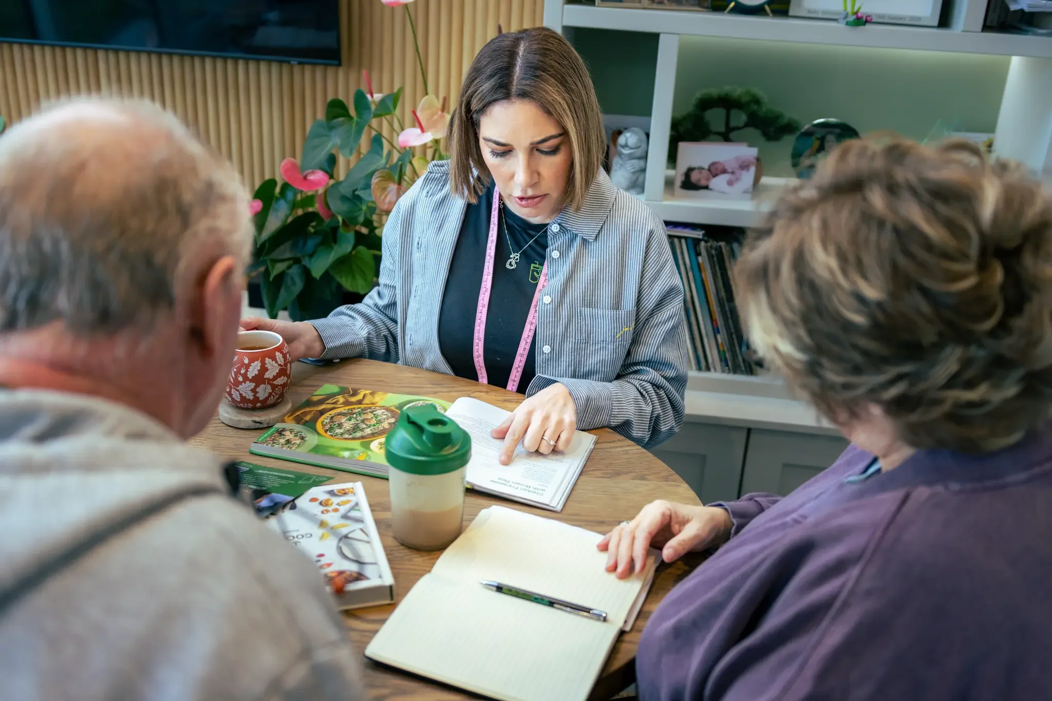 A woman explains something in a book to two older adults seated at a table. The table has a notebook, a pen, a magazine, a colorful food guide, a cup, and a protein shake on it. There are plants and bookshelves in the background.