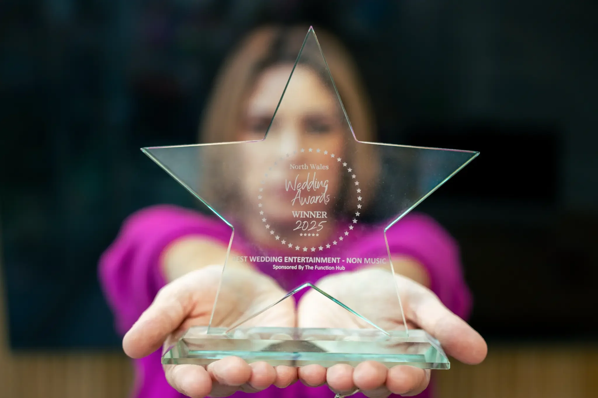 A woman in a purple shirt holding a transparent star-shaped award with inscriptions for the North Wales Wedding Awards 2025, in front of her face, with a blurred background.