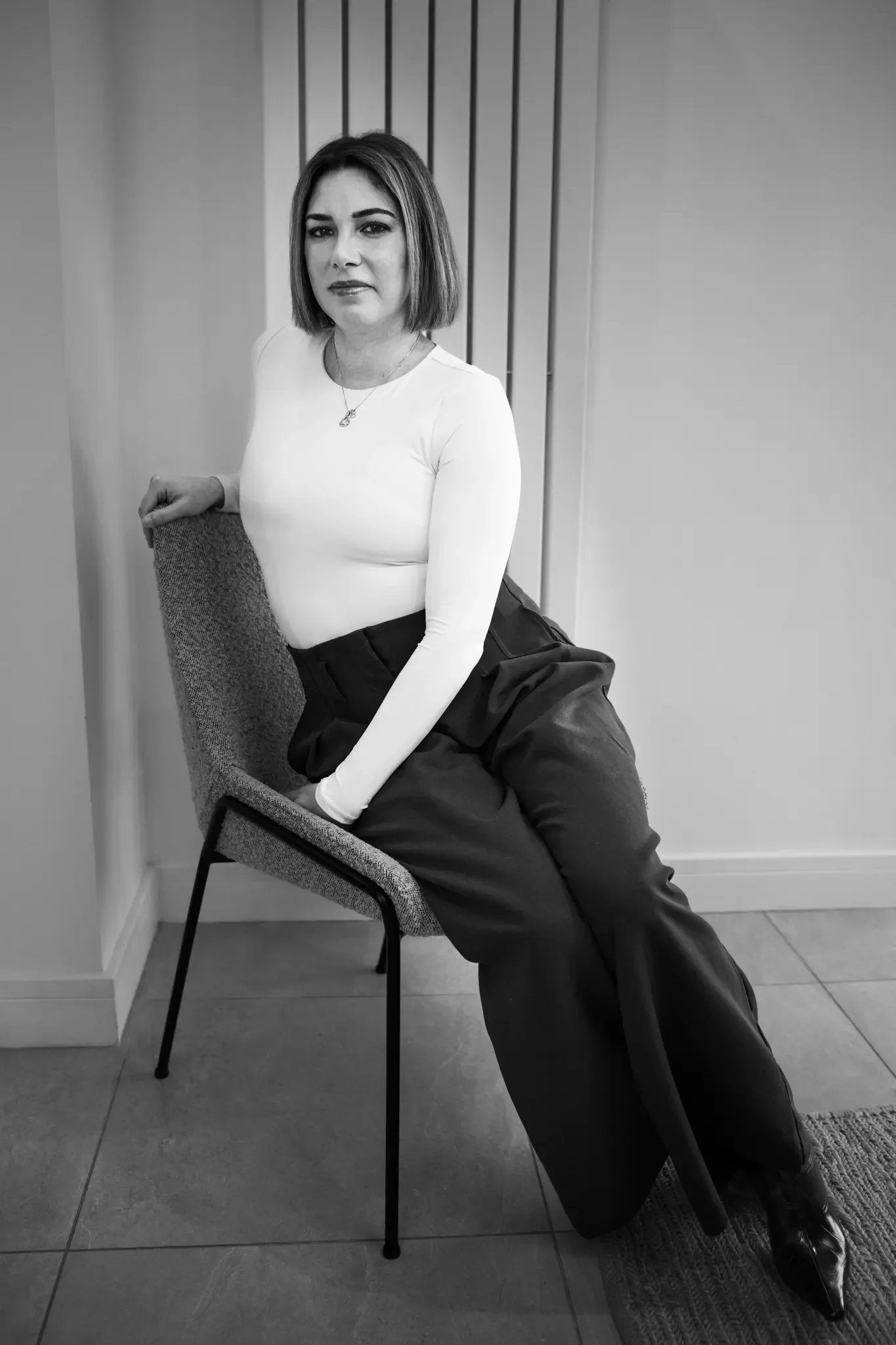 A woman with shoulder-length hair, wearing a white long-sleeve top, black pants, and shiny black shoes, sitting sideways on a chair with one hand on the backrest, in an indoor setting.