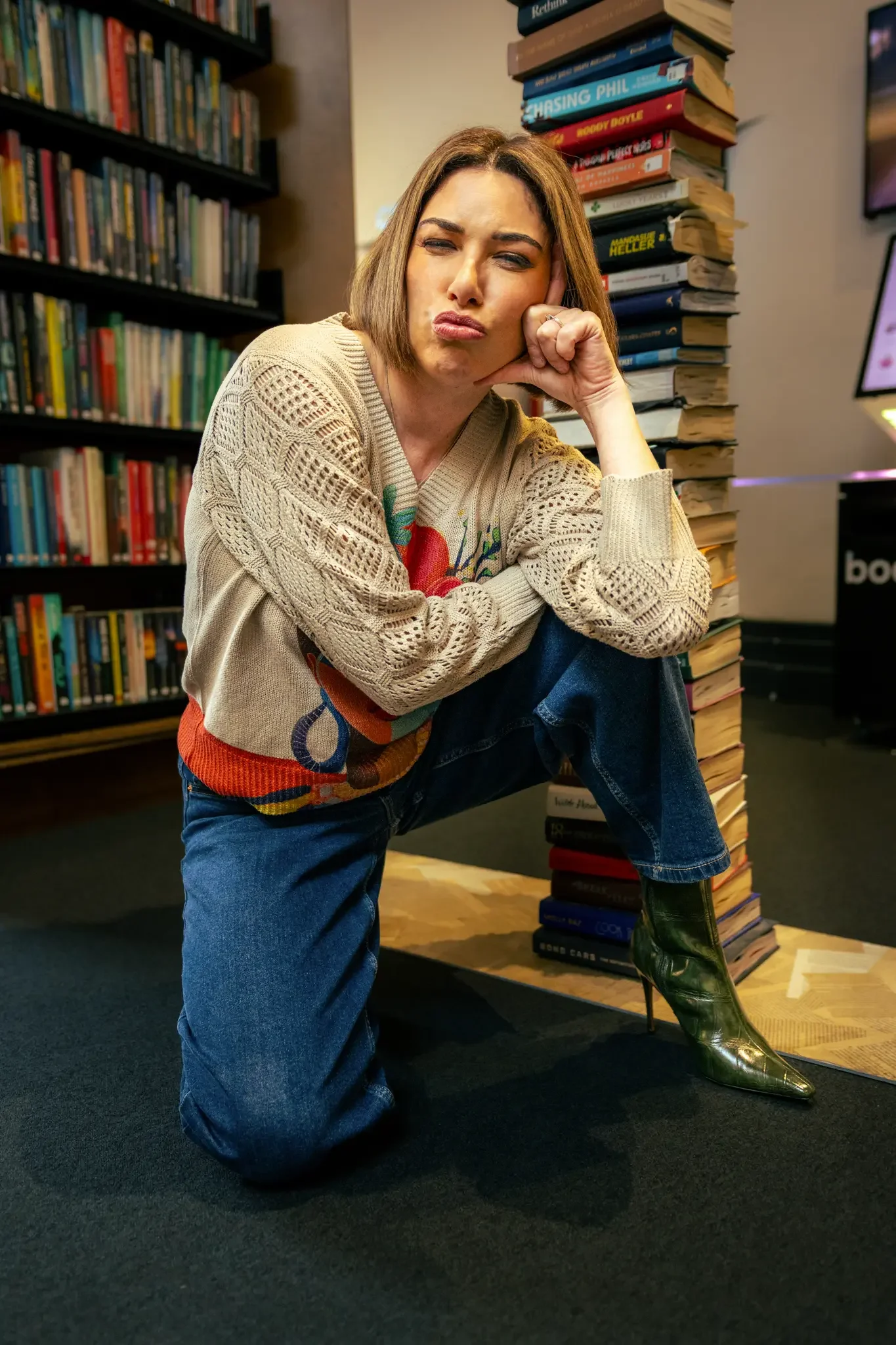 A woman with shoulder-length brown hair, wearing a knitted sweater with colorful embroidery, blue jeans, and shiny green high-heeled boots, sitting on one knee inside a bookstore near a tall stack of books and a bookshelf.