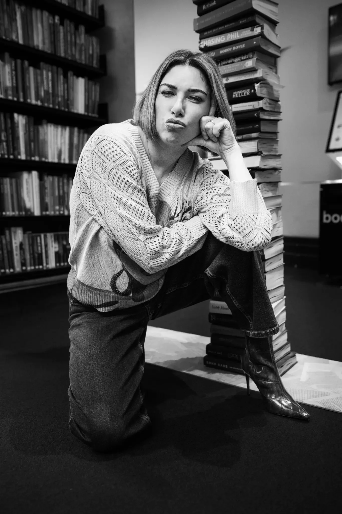 Black and white photo of a woman squatting on one knee in a bookstore, resting her chin on her fist, with a playful facial expression. She is wearing a patterned sweater, jeans, and high-heeled boots, with tall stacks of books around her.