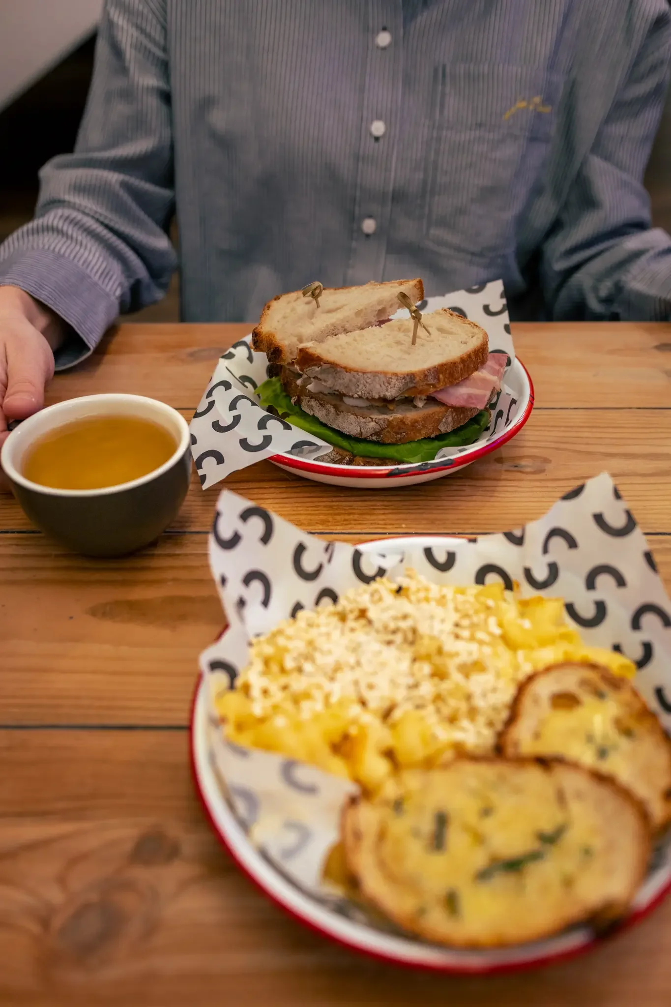 A person wearing a blue striped shirt sitting at a wooden table with a bowl of soup and two plates of food. The first plate has a sandwich with sliced bread, turkey, ham, and lettuce. The second plate contains macaroni and cheese and slices of toaste