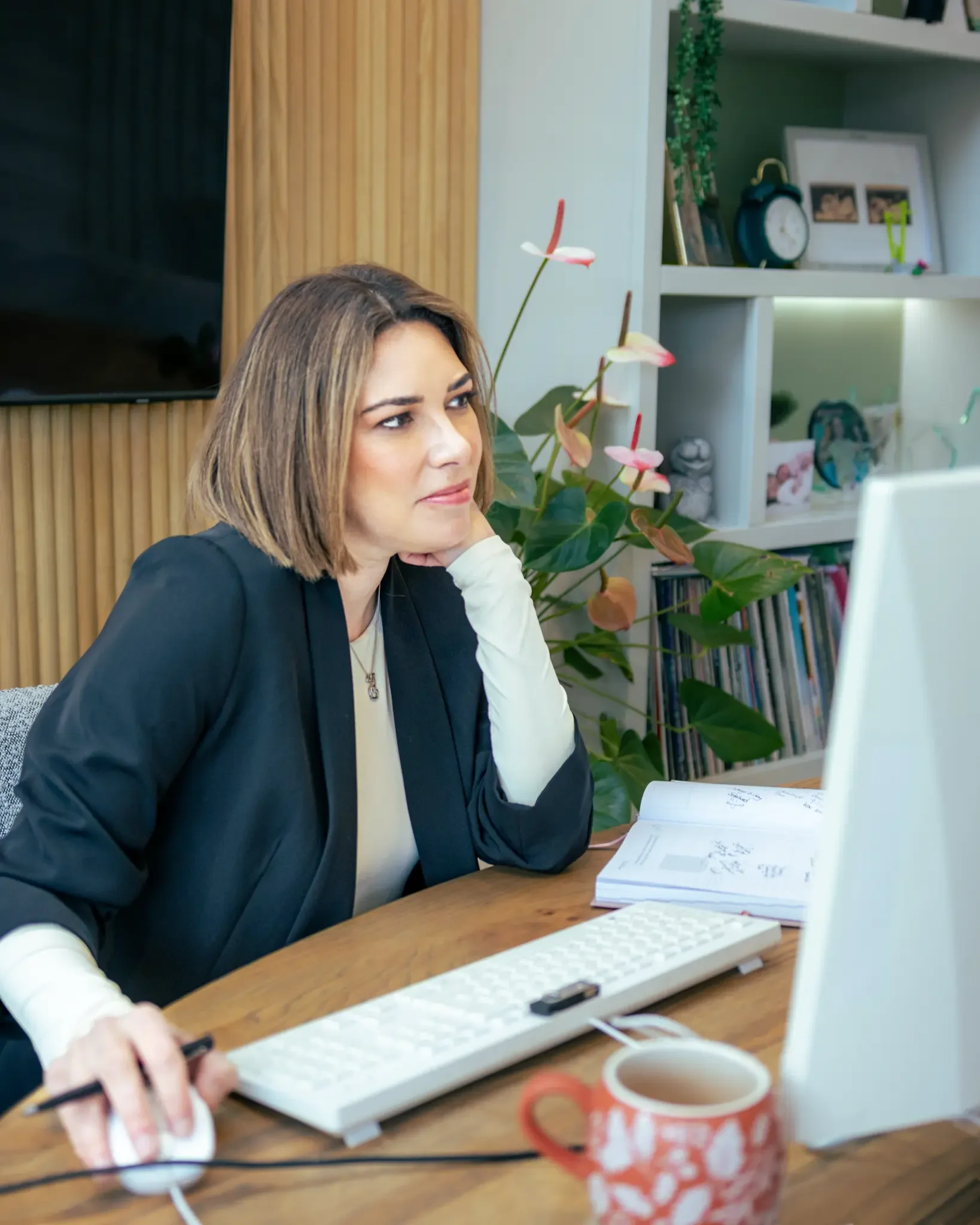 Woman working at a desk with a computer, open book, and coffee mug, in a home office with bookshelf and plants.