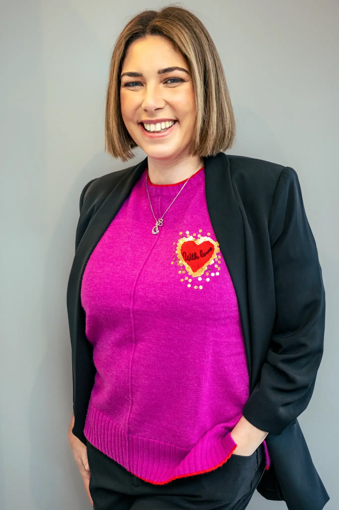 A woman with shoulder-length brown hair, smiling, wearing a black blazer over a bright pink sweater with a red and gold heart design that says 'With Love,' and a silver necklace.