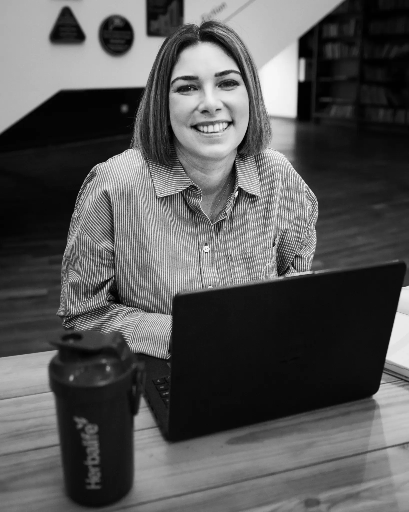 A woman smiling, sitting at a table with a laptop and a water bottle, in a room with shelves and wall decorations.