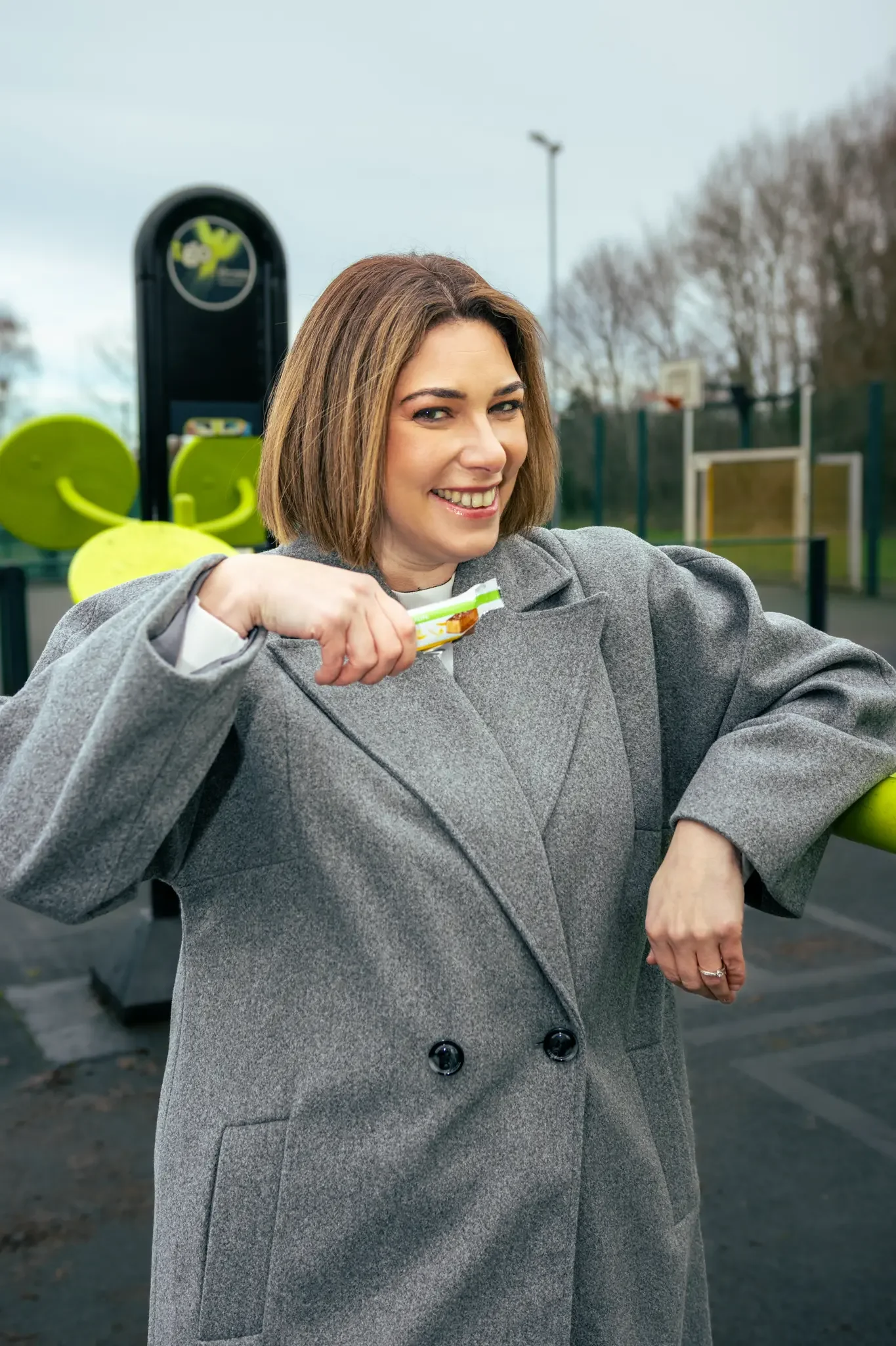 A woman in a gray coat smiling and holding a snack bar outdoors at a park or sports area.
