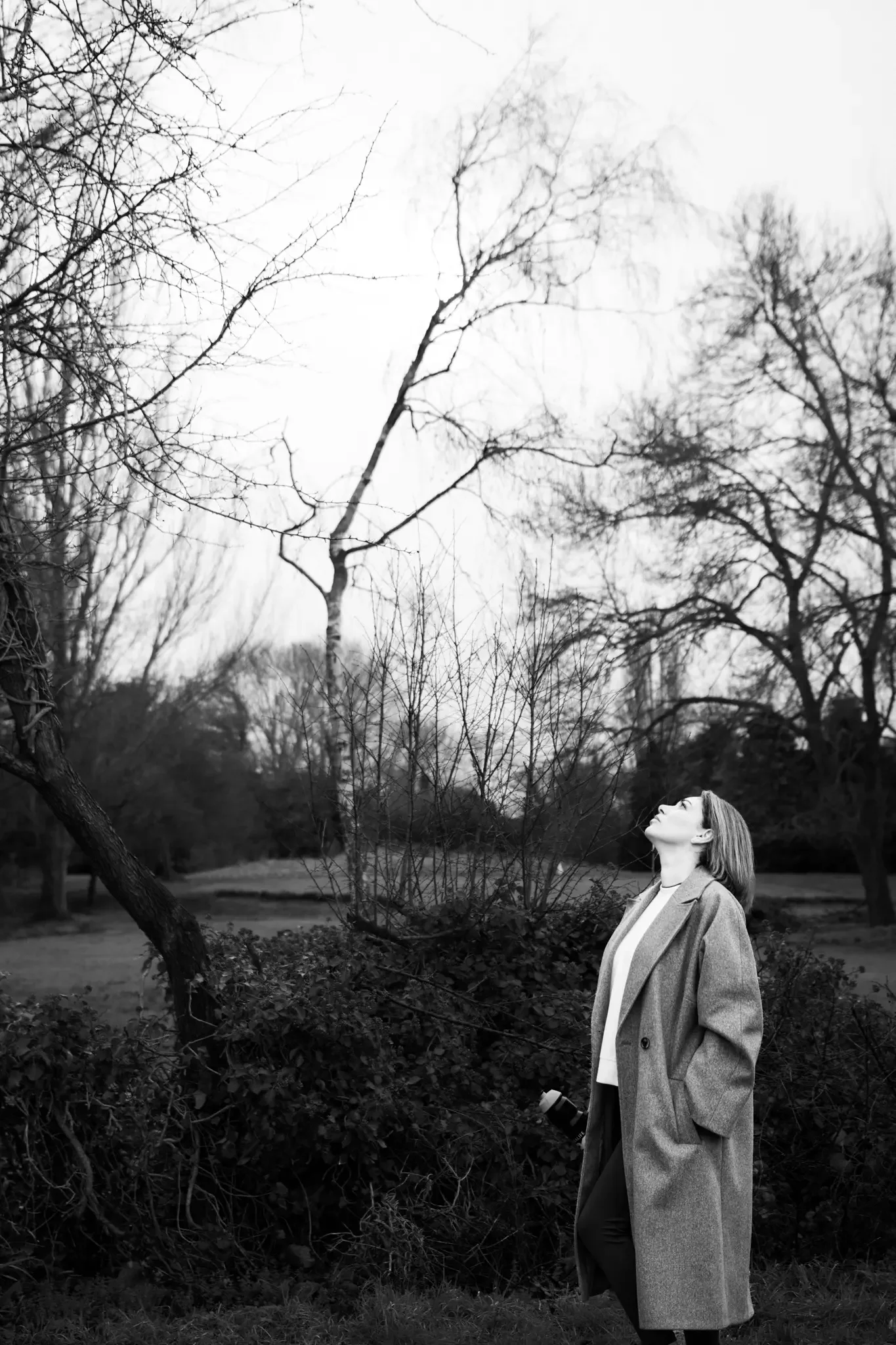 A woman in a coat standing outdoors in a park, looking up at the sky, with leafless trees in the background.