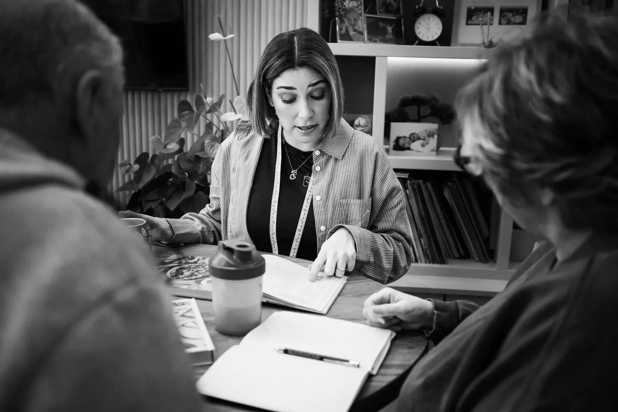 A woman discussing with two elderly people at a table, with books, a notebook, and a water bottle in a cozy room with bookshelves and plants.