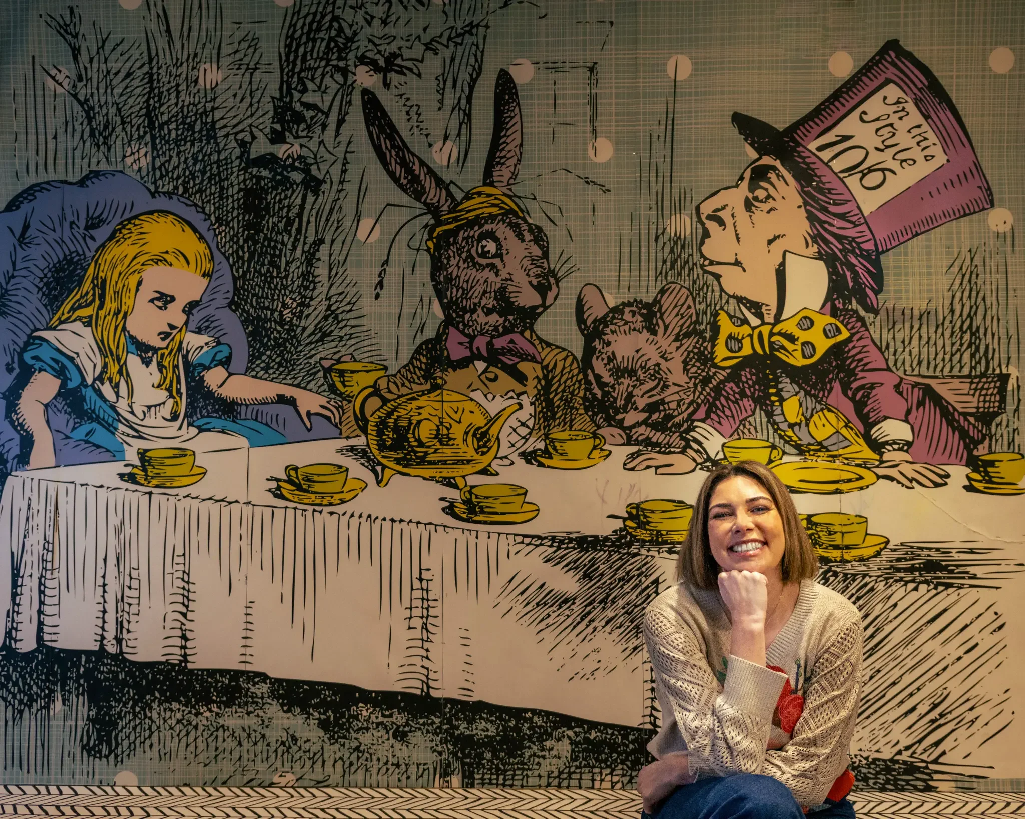 A woman with shoulder-length brown hair, smiling and sitting in front of a large cartoon wall art depicting Alice in Wonderland's Mad Hatter's tea party scene.