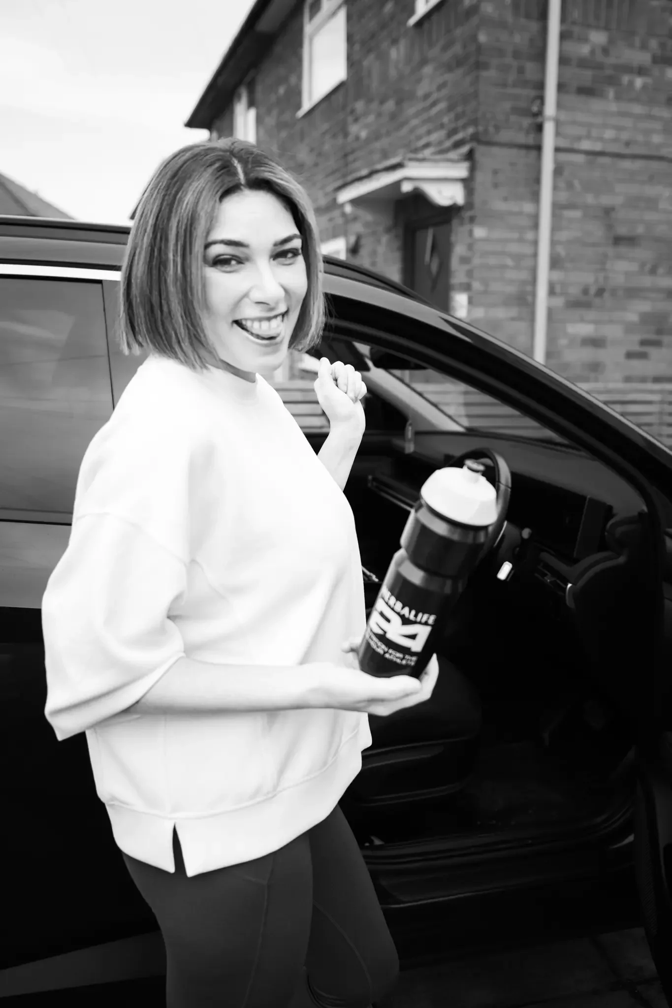 A woman with shoulder-length hair, smiling and holding a NealCalorie shaker bottle, standing beside a car with the driver’s side door open, in a residential neighborhood.
