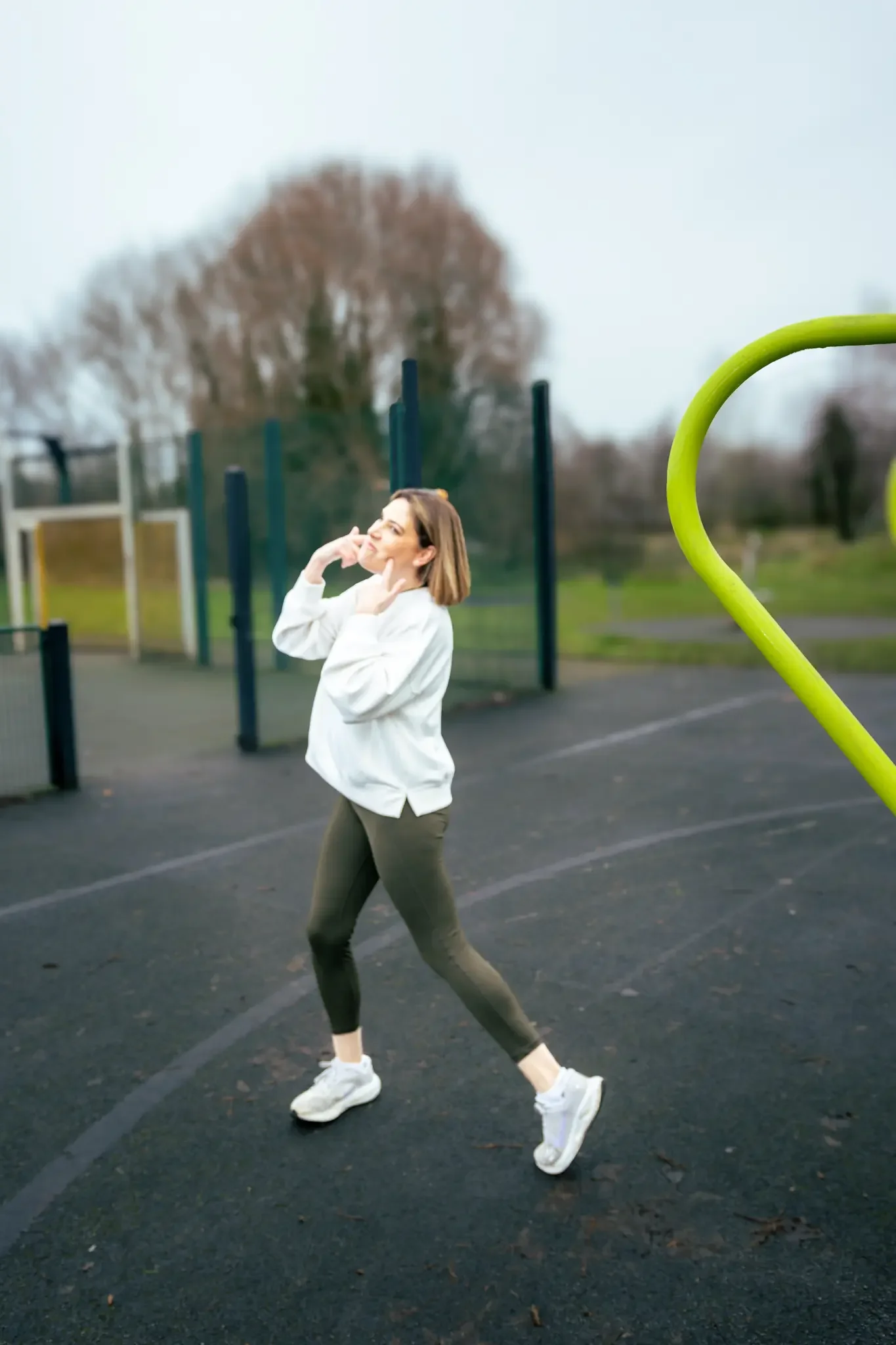 A woman in workout clothes posing outdoors in a park with an outdoor gym area visible in the background.
