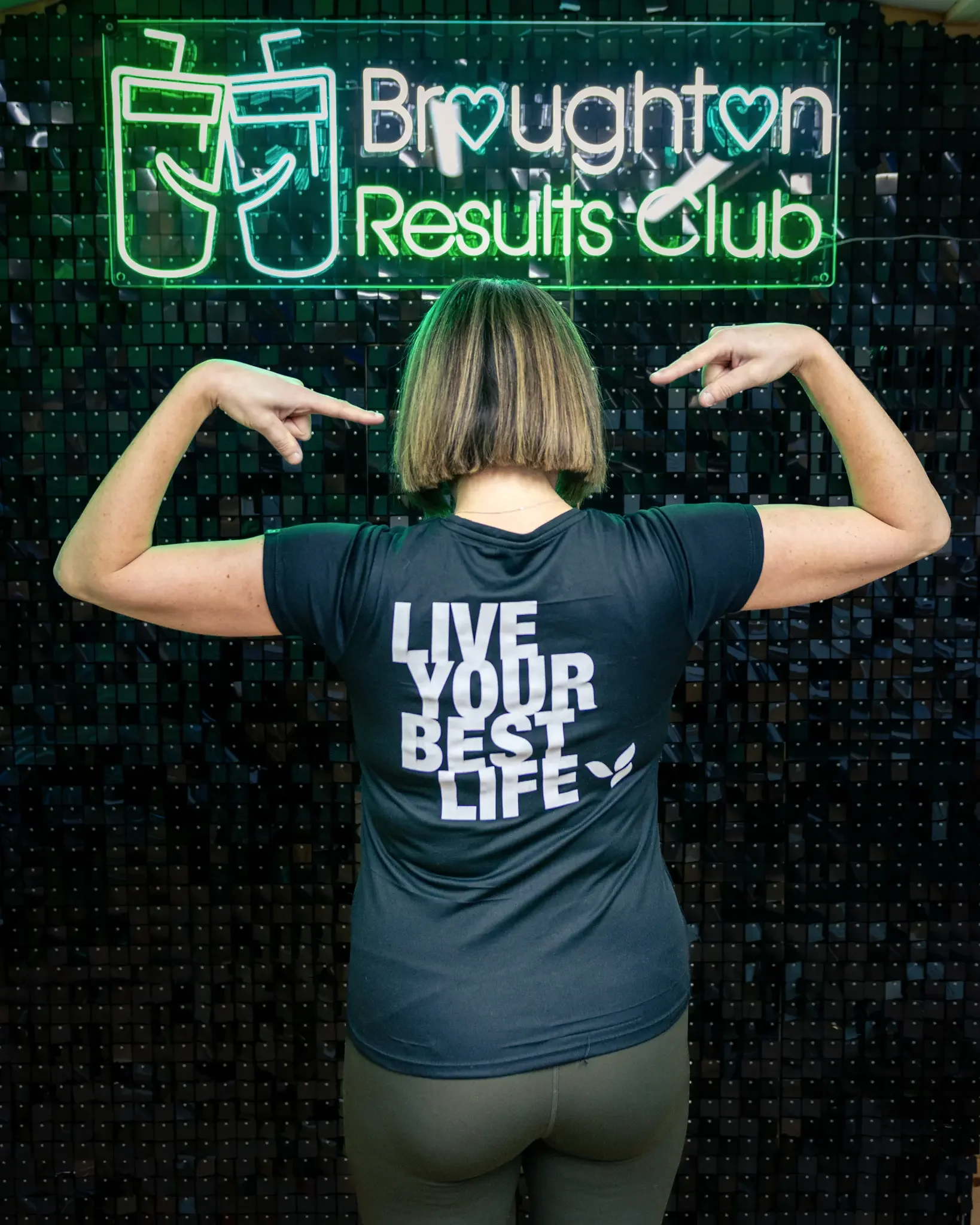 A woman with shoulder-length hair flexing her biceps in front of a black wall with a neon sign reading 'Brughton Results Club' and an illustration of two glasses of beer. She is wearing a dark t-shirt with the slogan 'LIVE YOUR BEST LIFE' on the back